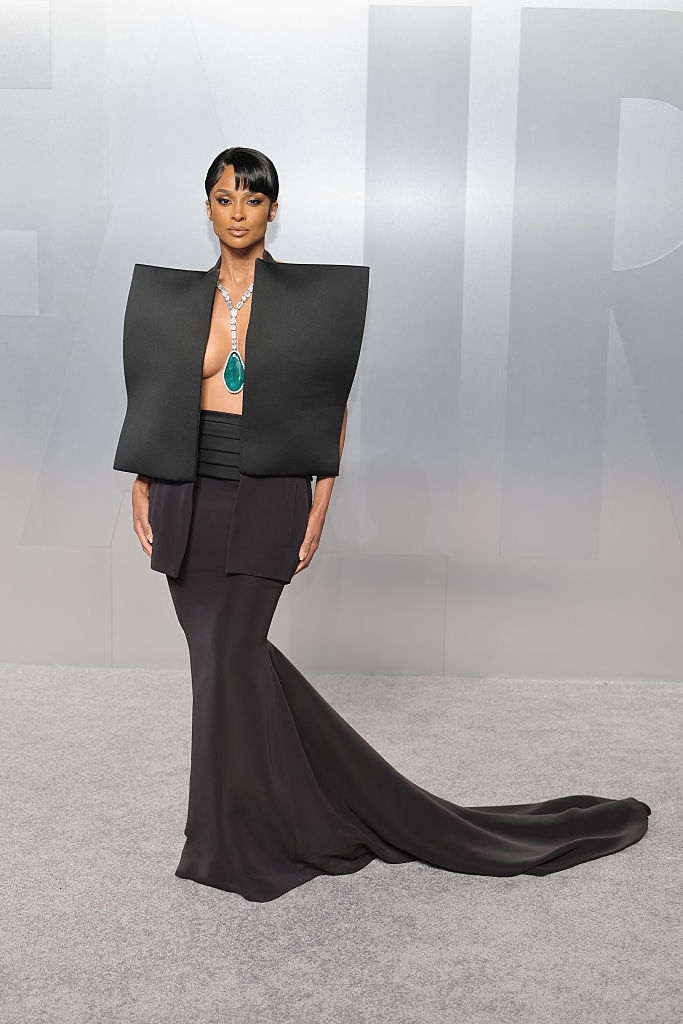 Person connected reddish carpet successful bold gown with geometric shoulders and mermaid skirt, wearing connection necklace with ample greenish gem