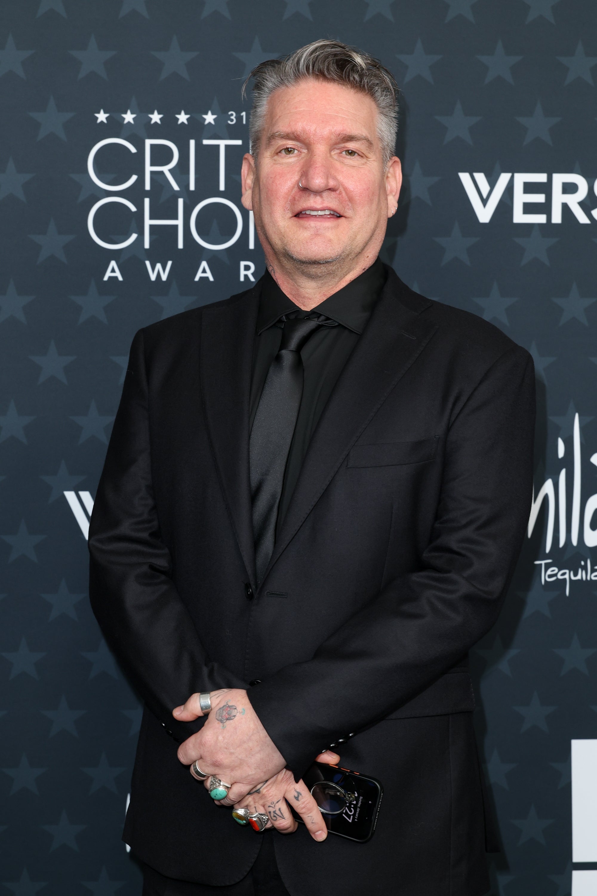 Person i en skreddersydd svart dress på en rød løper på en prisbegivenhet, poserer med et rolig uttrykk og holder en telefon