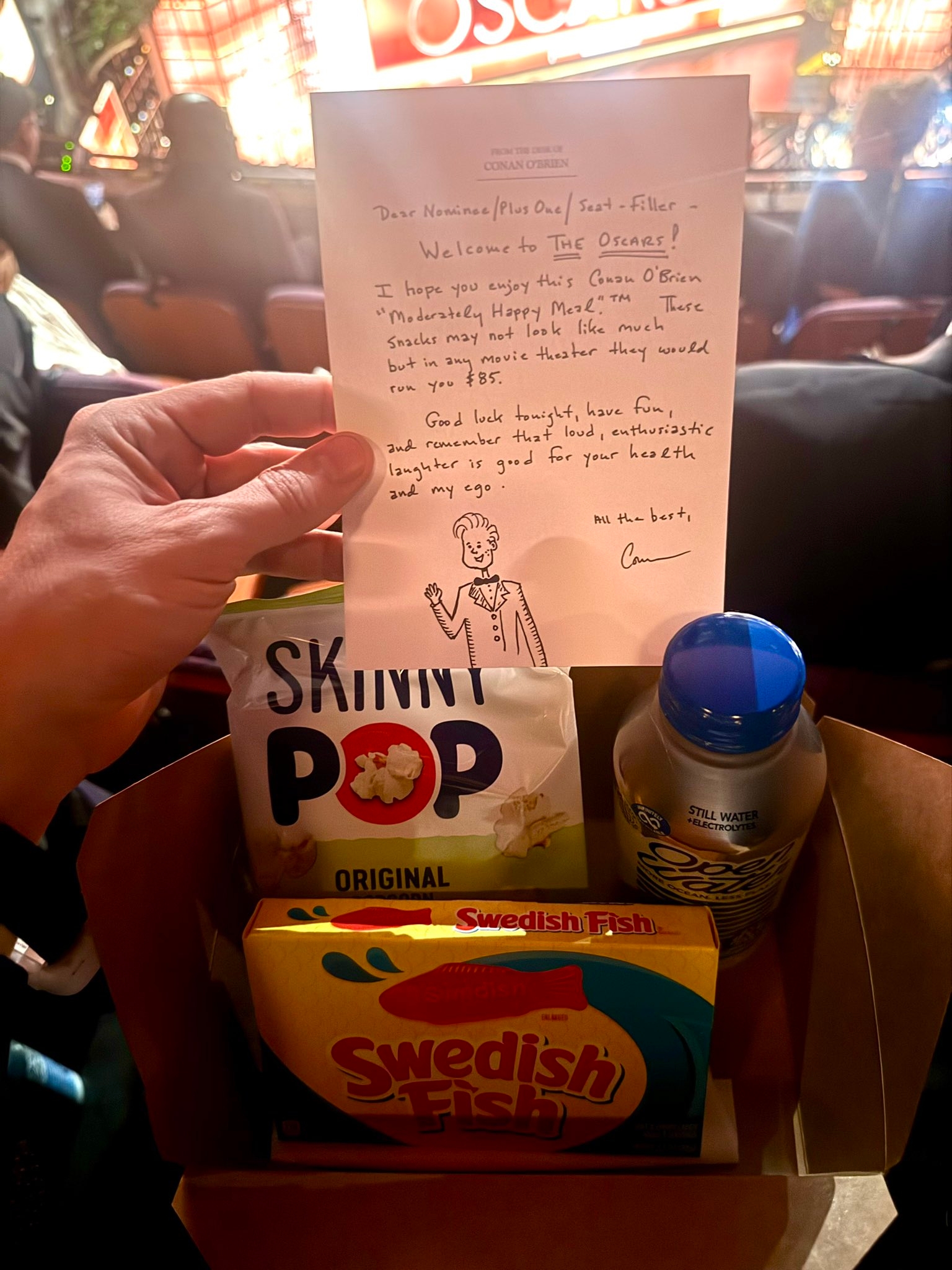 Hand holding a insubstantial container filled with snacks and a handwritten enactment welcoming attendees to an Oscars event