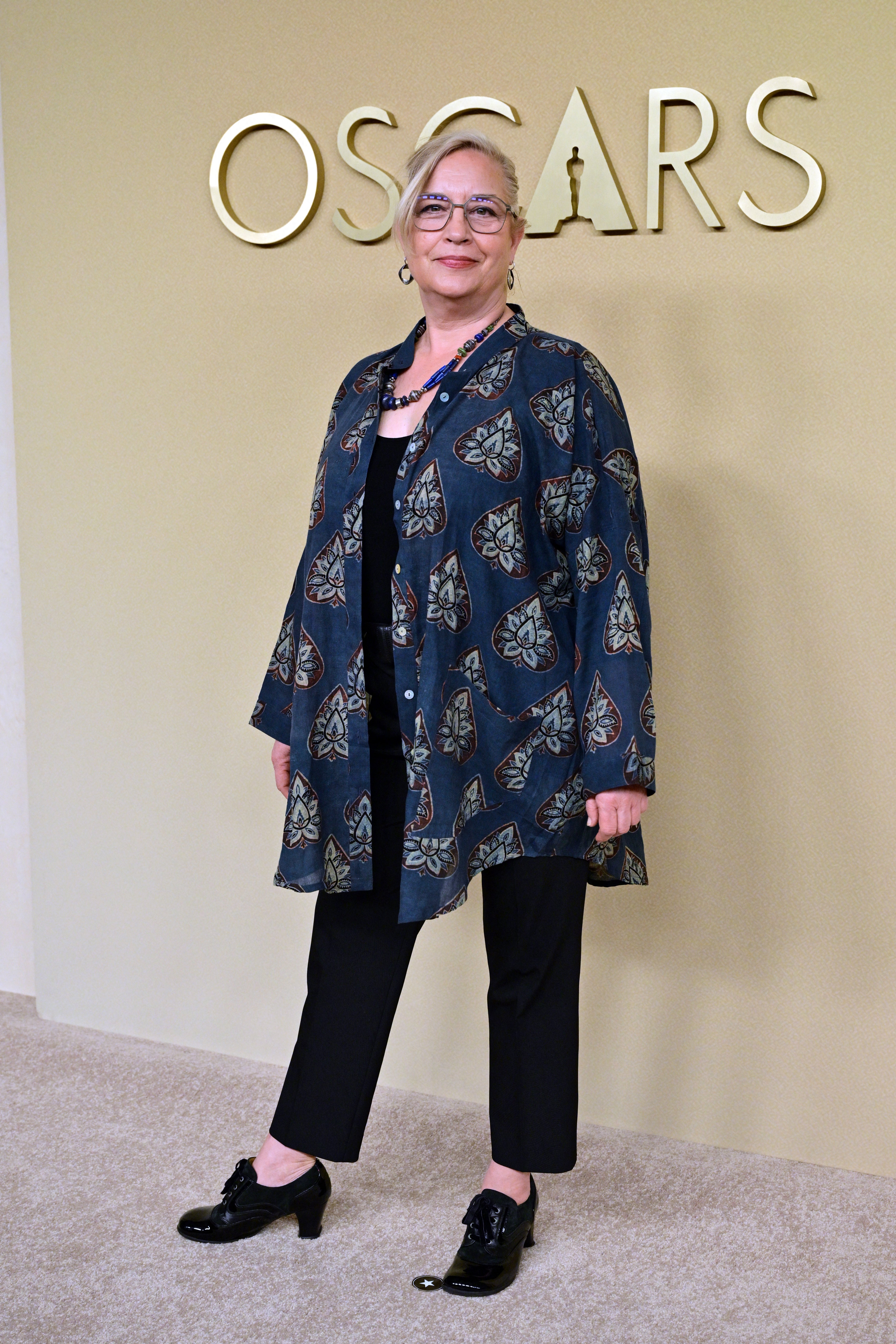 Personen står på Oscar-arrangementet, iført en mønstret lang skjorte, svarte bukser og sko, utstyrt med et halskjede og briller