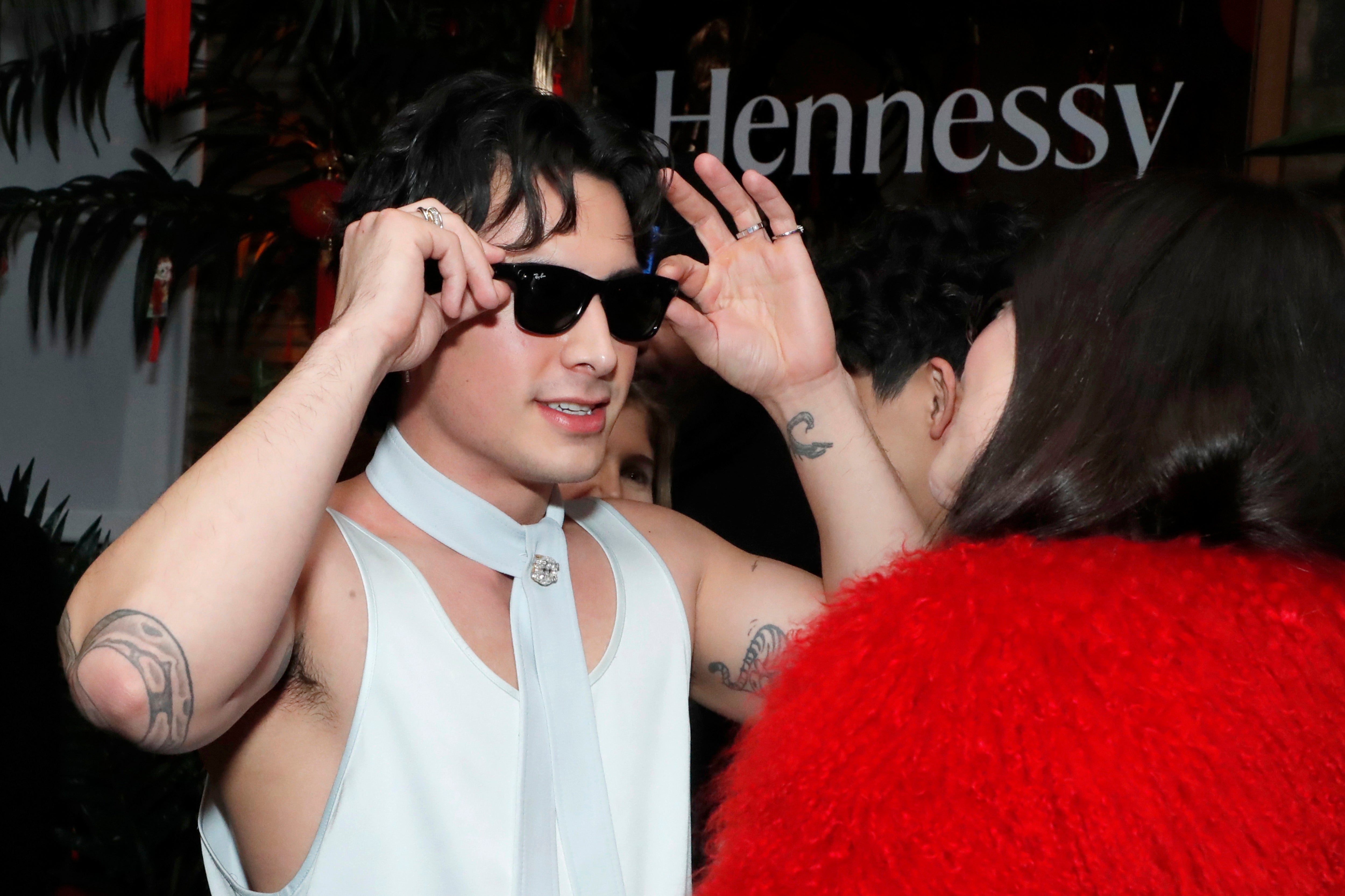 Person som justerer solbriller på et arrangement, har på seg en ermeløs topp med skjerftilbehør, snakker med noen i en dristig pelsjakke