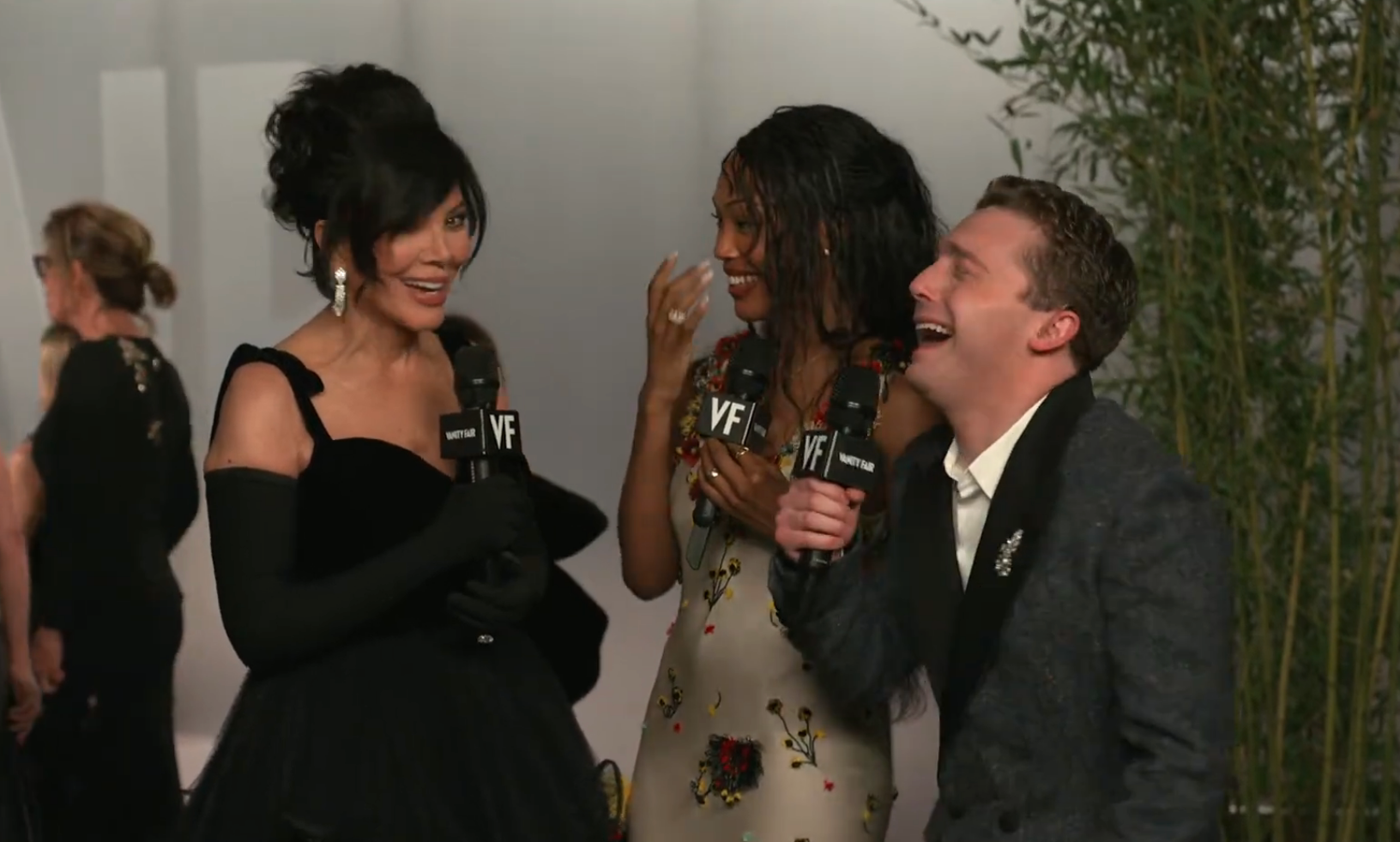 Three radical   smiling and holding microphones astatine  an event. One successful  a achromatic  gown and gloves, different  successful  a floral dress, the 3rd  successful  a acheronian  suit