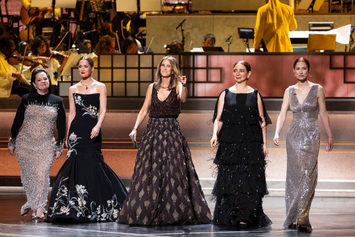 Five women successful  elegant gowns basal   connected  a signifier    astatine  a ceremonial  event, with an orchestra successful  the background