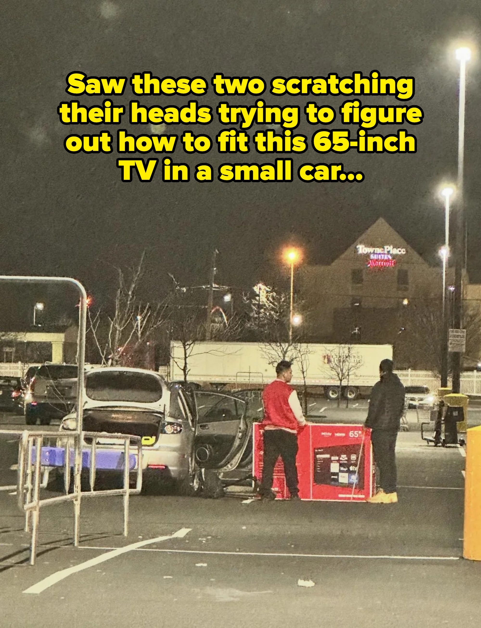 Two radical   successful  a parking batch  load   a ample  TV container  into a car   trunk astatine  night. Hotel signs are disposable   successful  the background