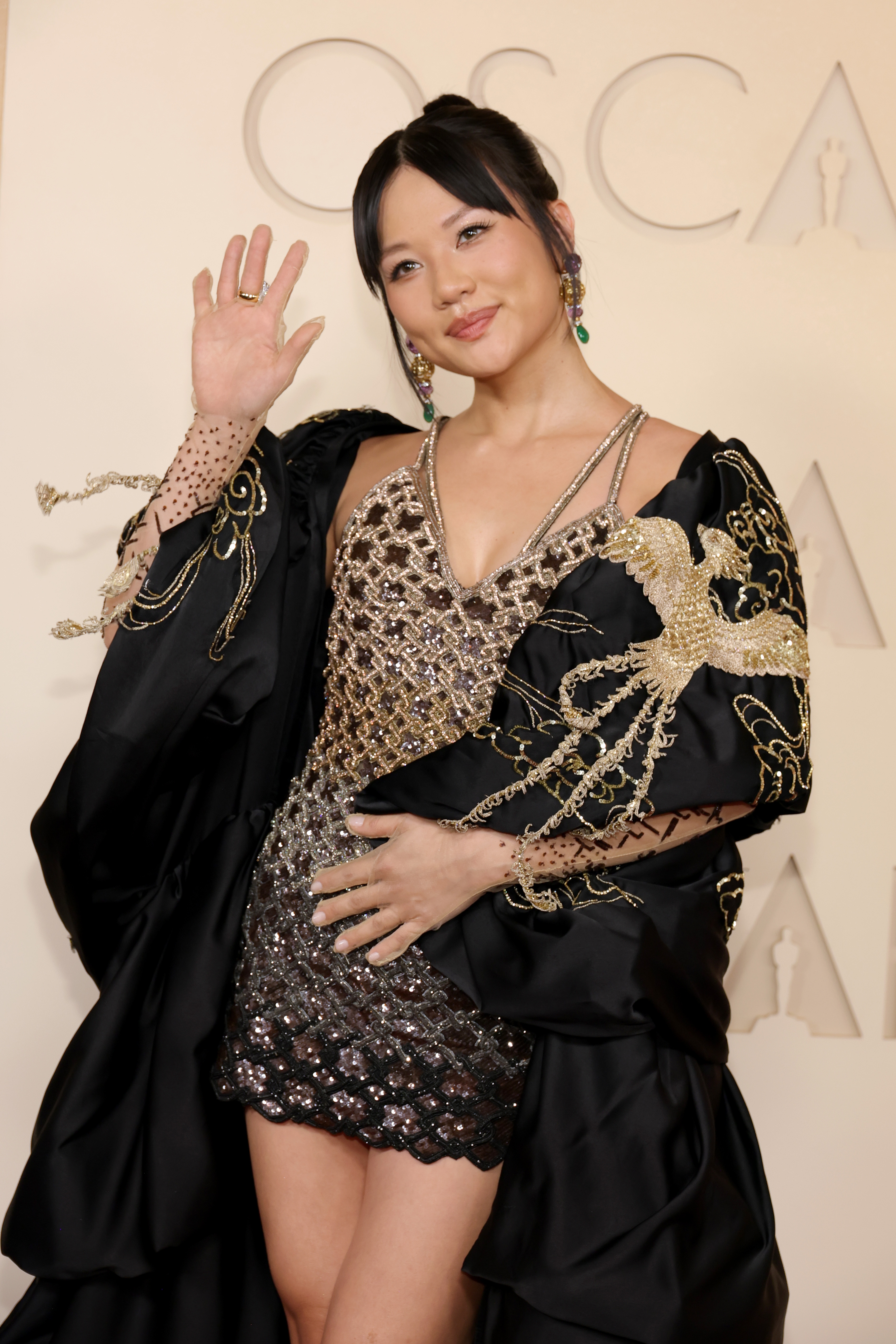 A pistillate   poses connected  a reddish  carpet successful  an embellished gown with large, elaborate  sleeves, waving and smiling