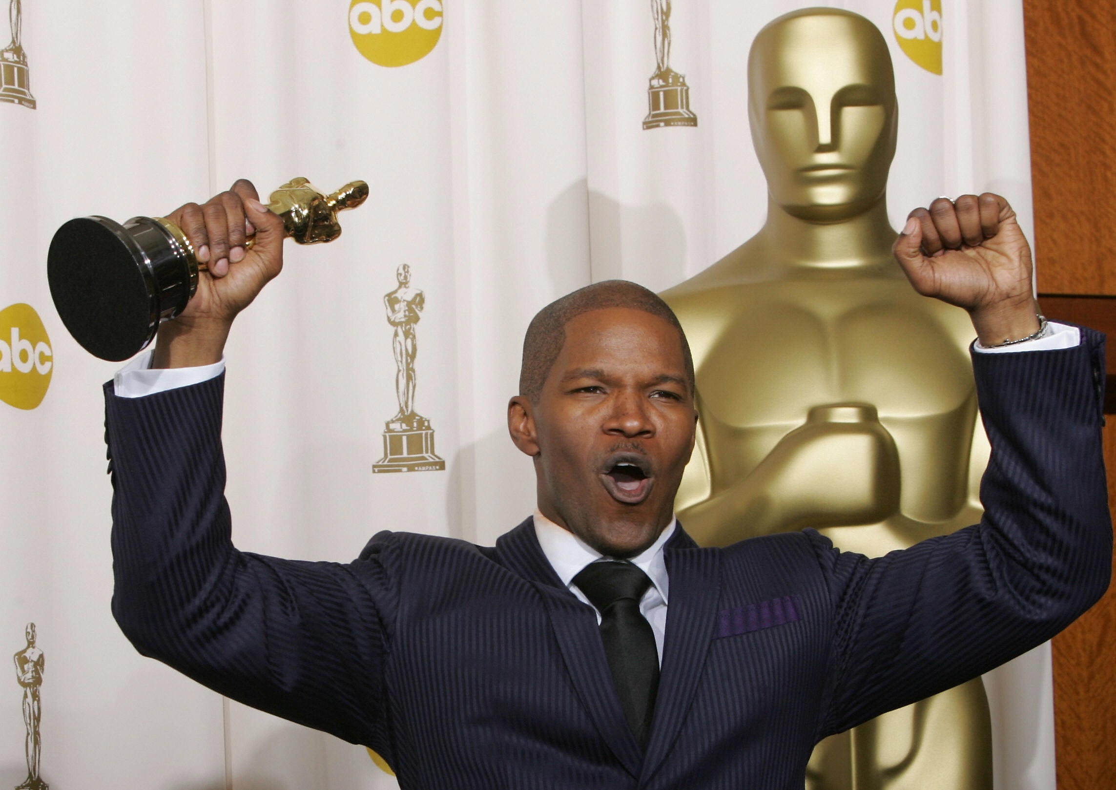 Person i dress feirer med et Oscar-trofé i hånden, stående foran en stor Oscar-statue