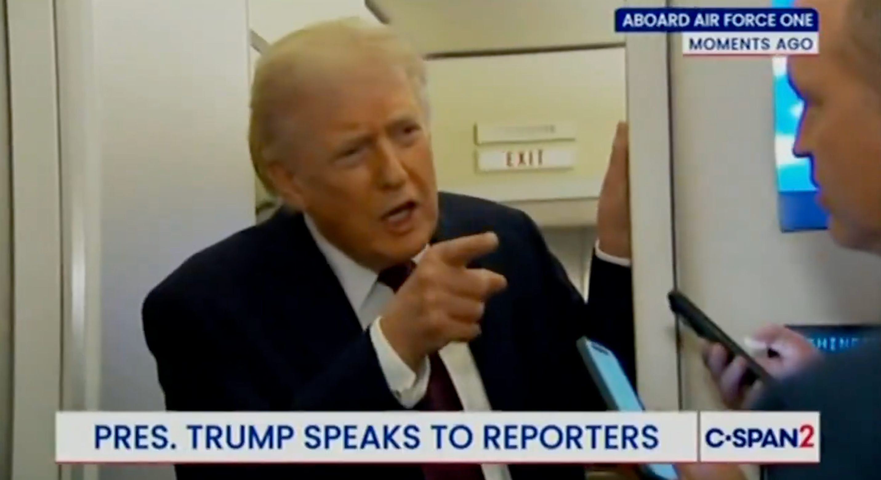 Person som snakker med journalister ombord på Air Force One, gestikulerer mens han deltar i en samtale. Nyhetsbanneret sier, 