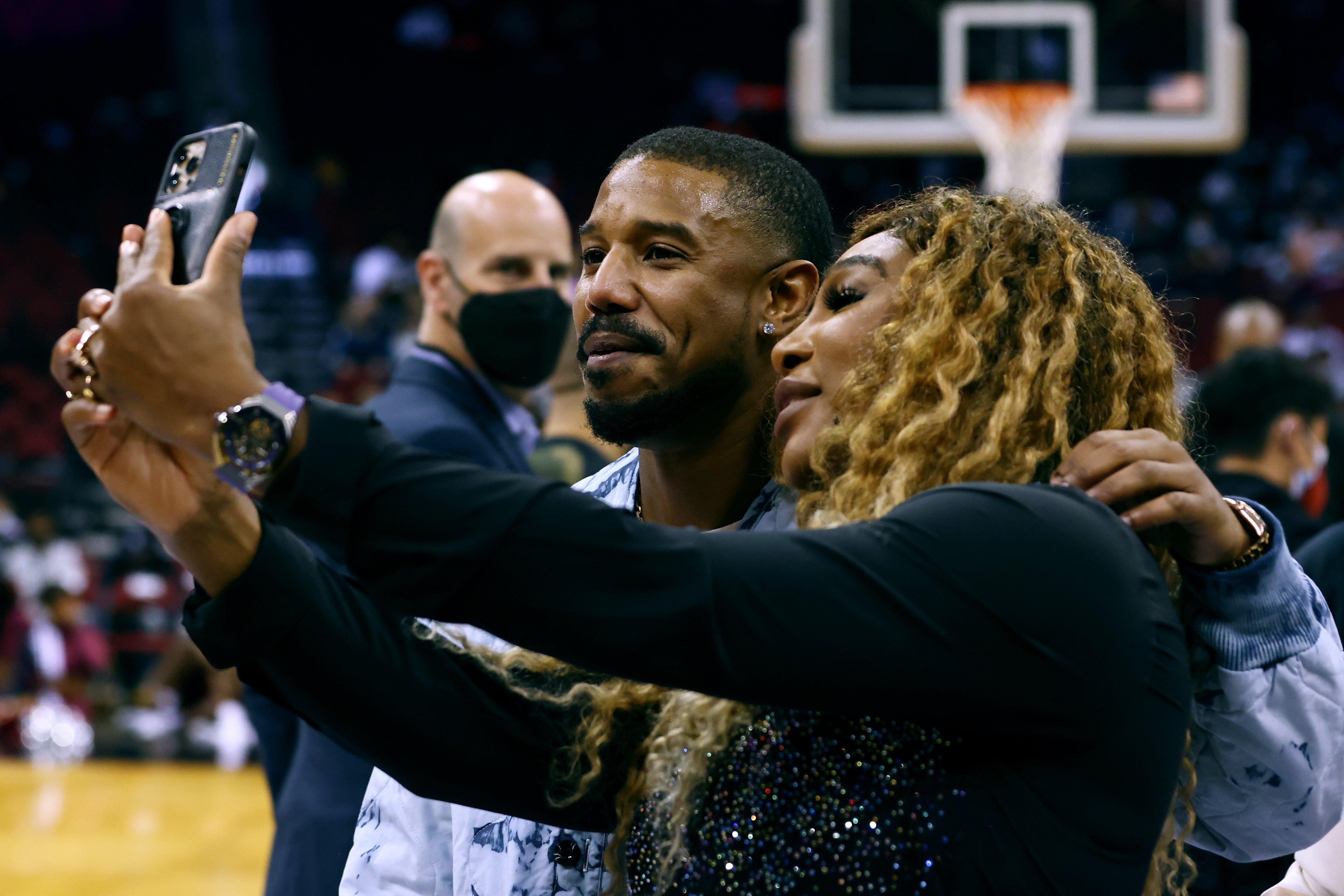 To personer tar en selfie ved en basketballkamp. Den ene har på seg en mønstret jakke, og den andre har langt krøllete hår og en glitrende topp