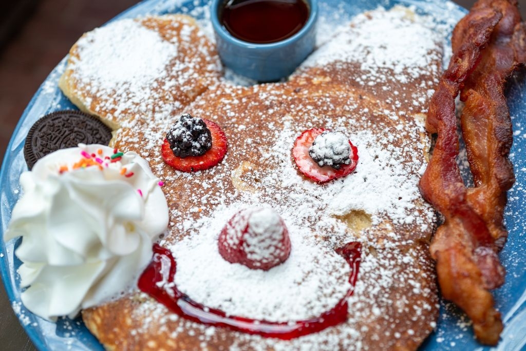 Pancake shaped similar  a bear's face, topped with effect   and whipped cream, with bacon strips connected  the side