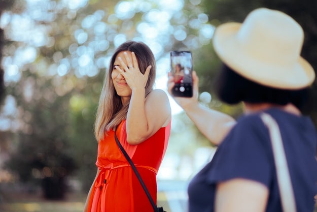 Person successful  absorption   playfully hiding look   from a camera held by different  idiosyncratic   wearing a hat. Casual outdoor setting