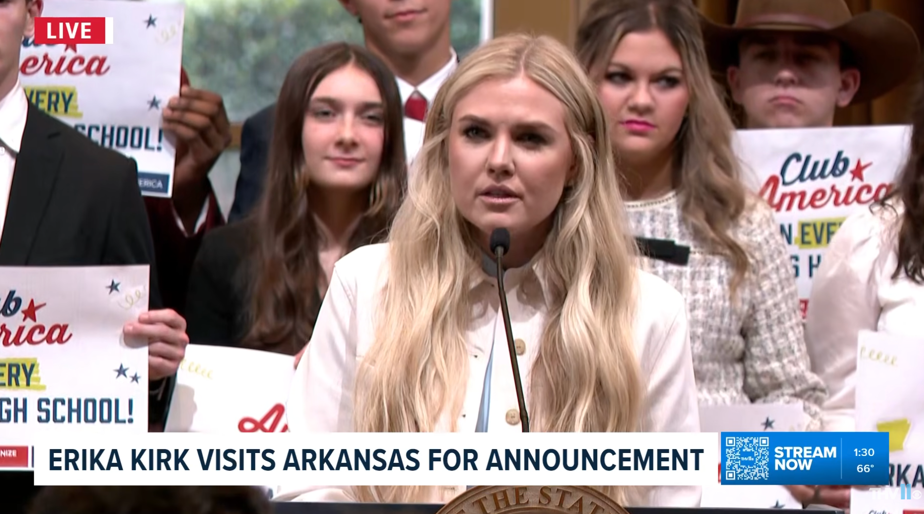 En kvinne taler ved et podium omgitt av folk som holder skilt med teksten 