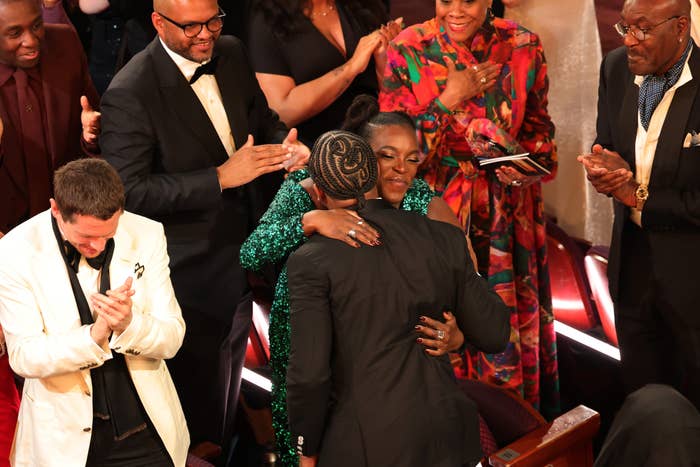 A idiosyncratic   successful  a glamorous sequined formal  hugs idiosyncratic    successful  a suit   astatine  a ceremonial  event, surrounded by applauding attendees successful  evening attire