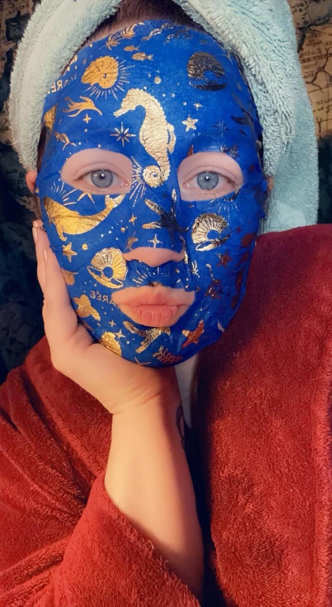 Person wearing a celestial-themed face mask and a towel on their head, wrapped in a plush robe, posing with a hand on their cheek