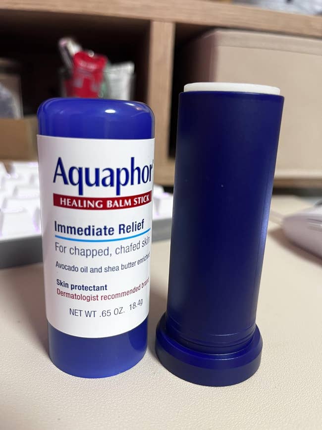 Aquaphor Healing Balm Stick and open cap on a desk, offering relief for chapped skin, featuring avocado oil and shea butter