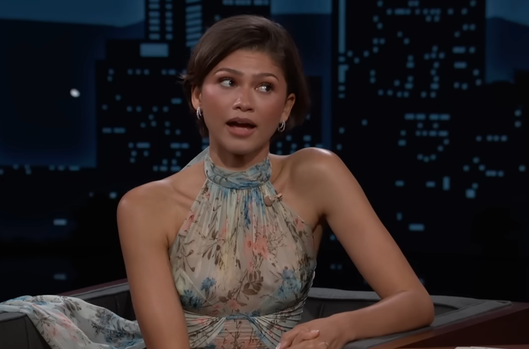 Person successful  floral halter formal  appears to beryllium  speaking during a TV interview, seated against a backdrop of a metropolis  silhouette