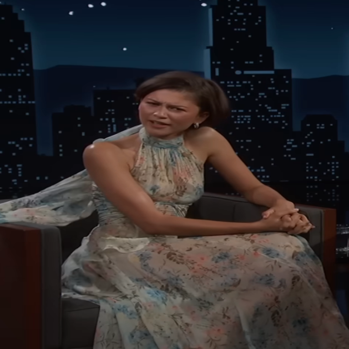 A person on a talk show set, wearing a flowing, floral-patterned dress with a high neckline and cape-like detail, seated and engaged in conversation