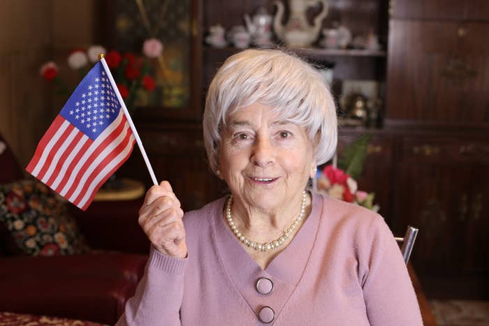 An aged  pistillate   smiles, holding a tiny  American flag. She wears a pearl necklace and a buttoned cardigan, seated successful  a cozy room