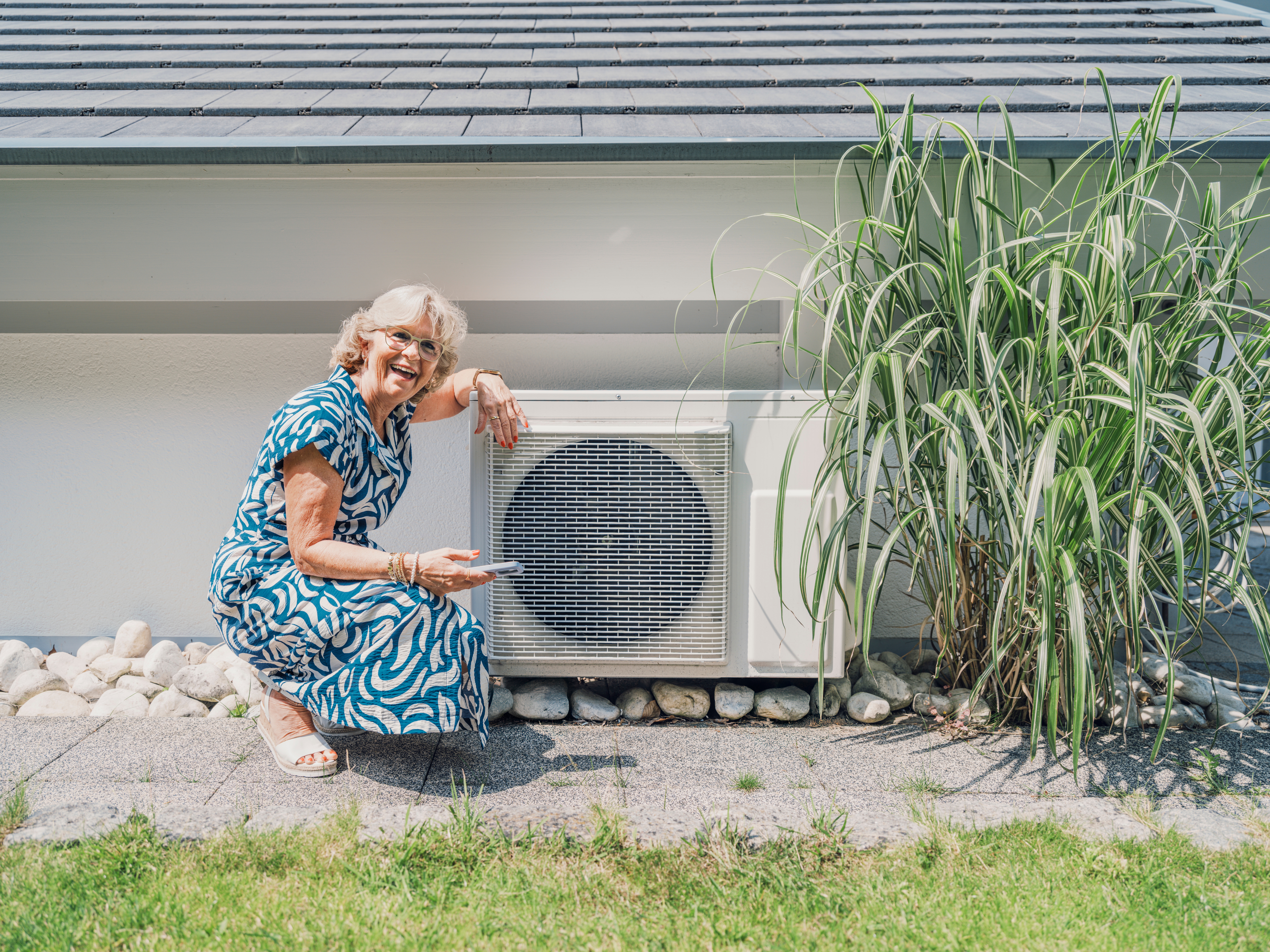An older pistillate   smiling, kneeling beside an outdoor aerial  conditioning unit, pointing astatine  it. She's wearing a patterned formal  and sunglasses