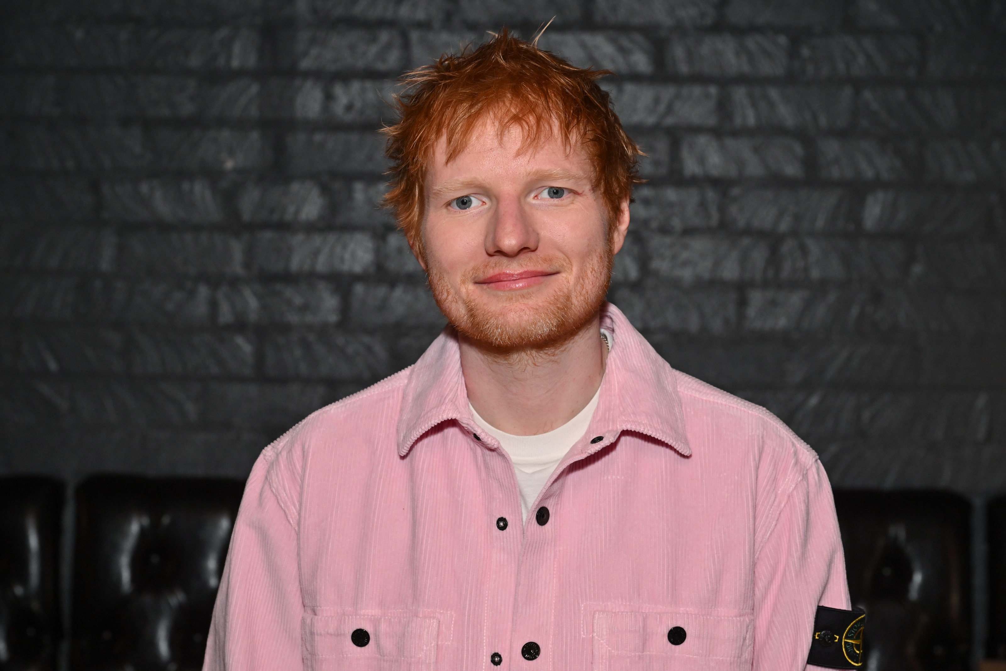 Ed Sheeran, with tousled hairsbreadth  wearing a casual button-up shirt, poses against a acheronian  ceramic  backdrop