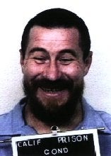 A man with a beard smiling while holding a sign below, possibly a mugshot