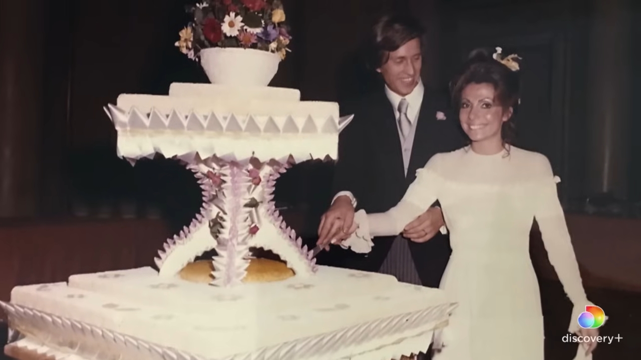 A mates smiles adjacent to a large, tiered wedding barroom decorated with intricate designs and topped with flowers