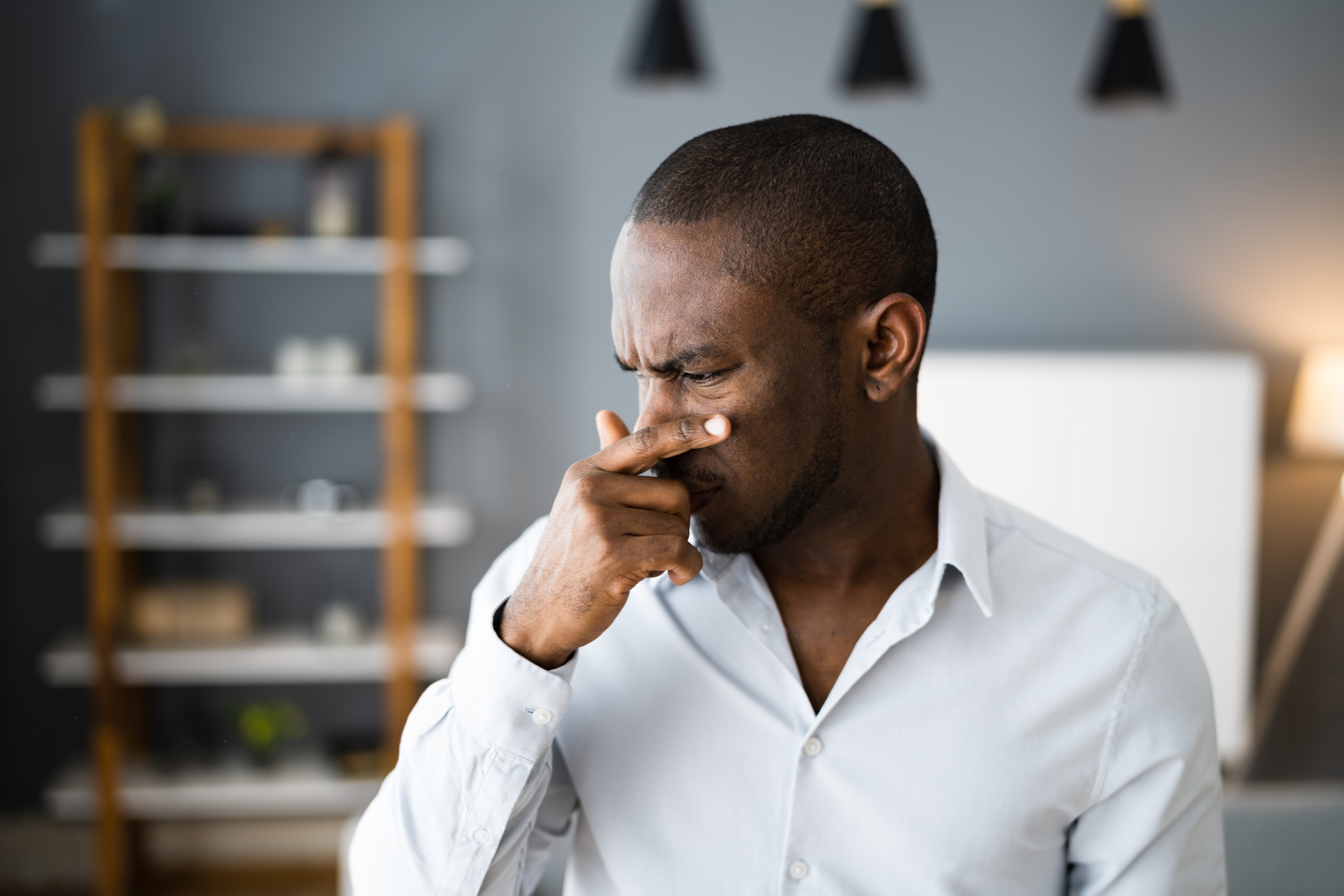 Man in a button-up shirt pinching his nose in discomfort, possibly due to an unpleasant smell, in a modern room setting
