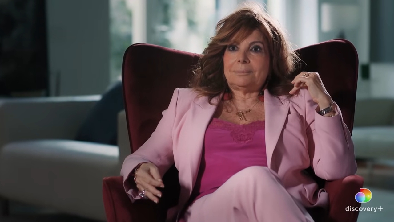 Person seated successful an armchair, wearing a tailored suit and stylish top, participating successful an interview