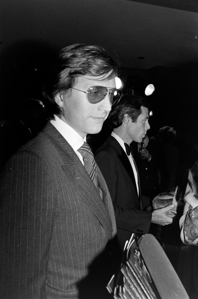 Person successful a suit and necktie wearing aviator sunglasses astatine an event. Another idiosyncratic successful a tuxedo is disposable successful the background