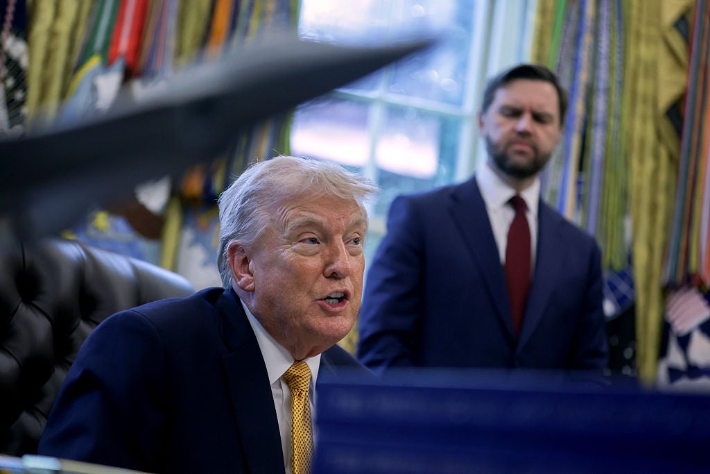 En person i dress sitter ved et skrivebord og snakker, med en annen person ved siden av dem. De er i kontormiljø med flagg i bakgrunnen