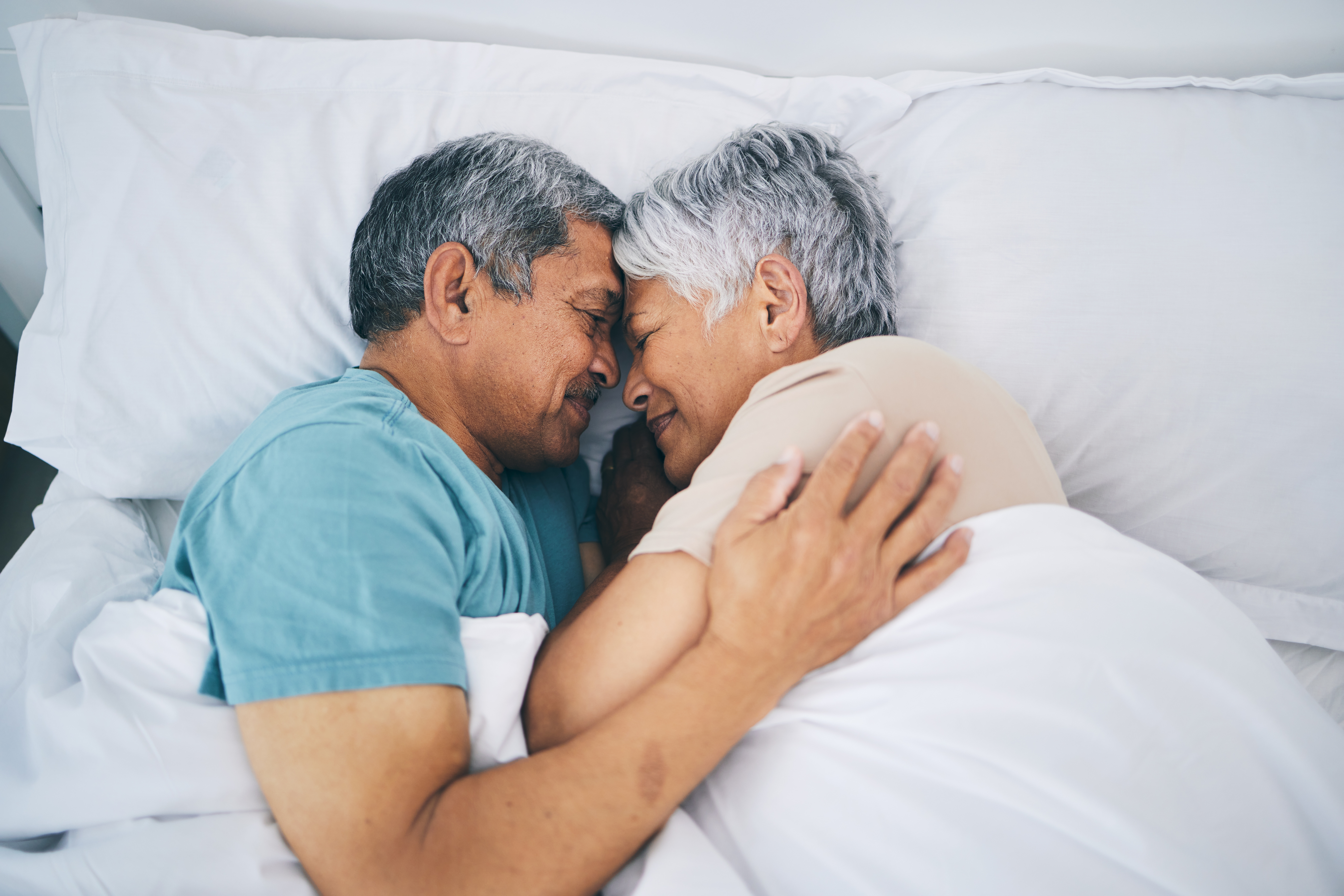 An elderly couple lovingly cuddles in bed, facing each other with gentle smiles, conveying warmth and intimacy