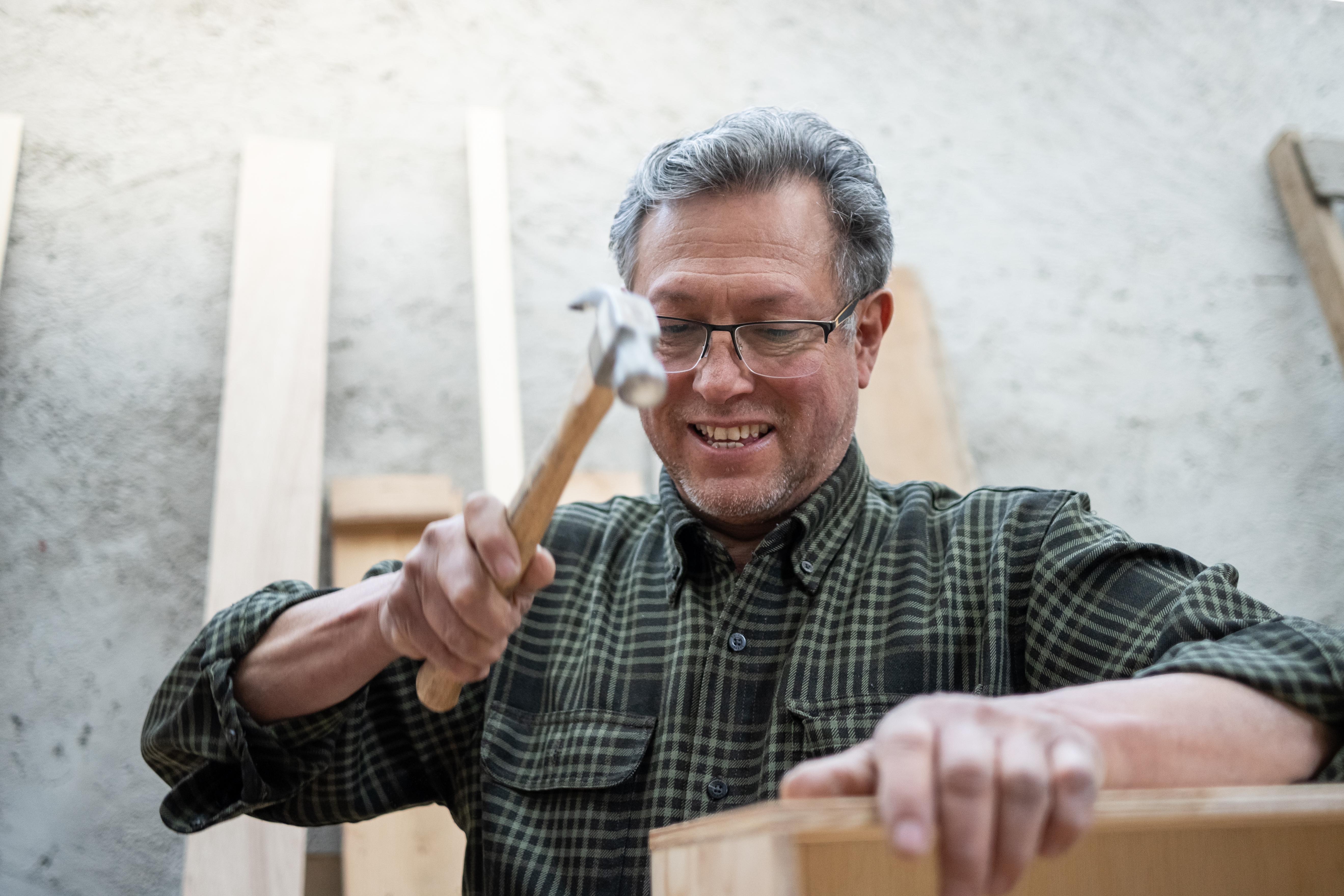 Man in plaid shirt smiling while hammering a nail into wood, surrounded by wooden planks