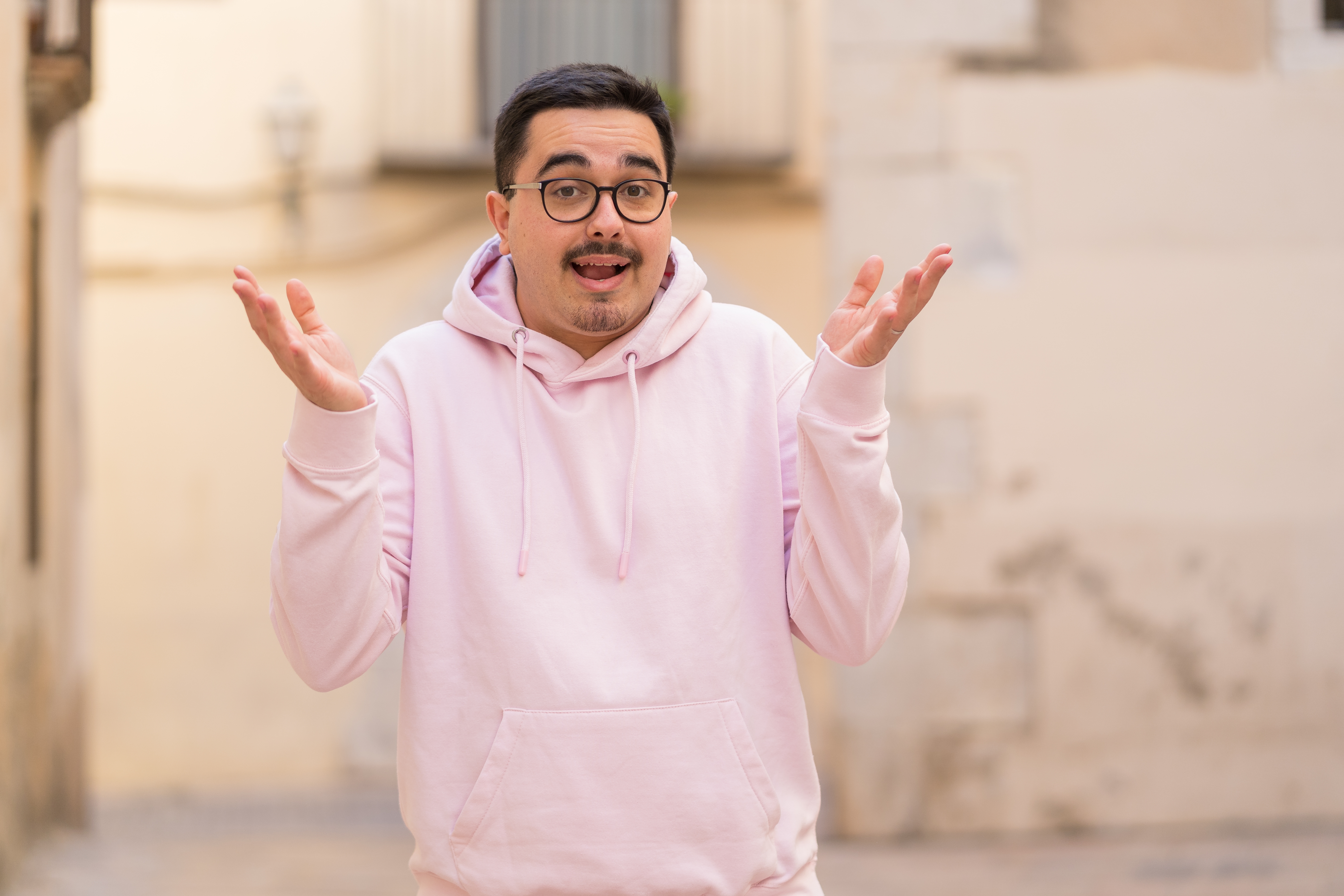 Person shrugging with hands up, wearing a casual hoodie and glasses, standing outside on a street