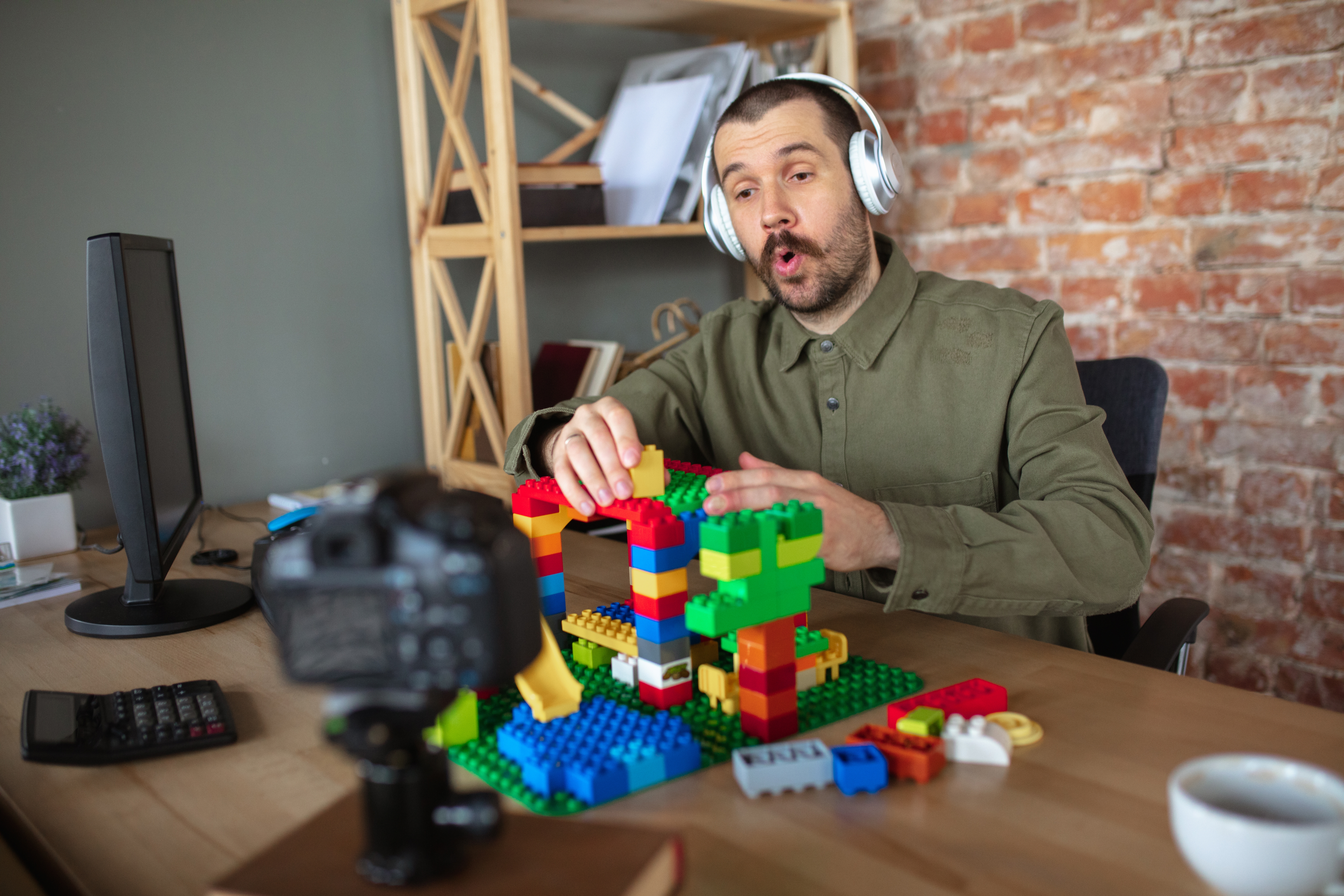 Person wearing headphones, engaged and expressive, builds with colorful blocks at a desk, while being recorded, in a casual home office setting
