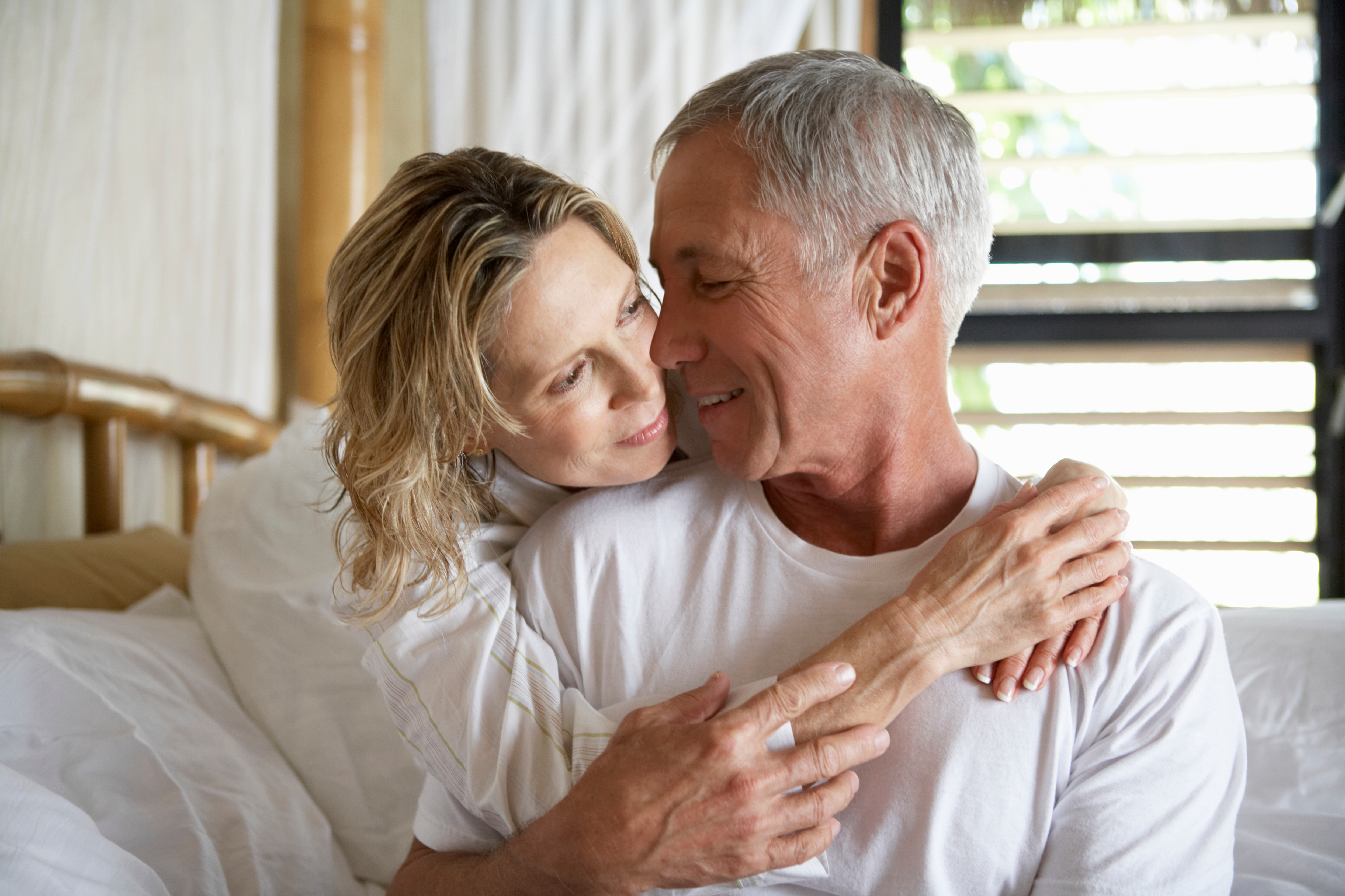 An older couple embraces warmly on a bed, looking into each other's eyes with affection and happiness