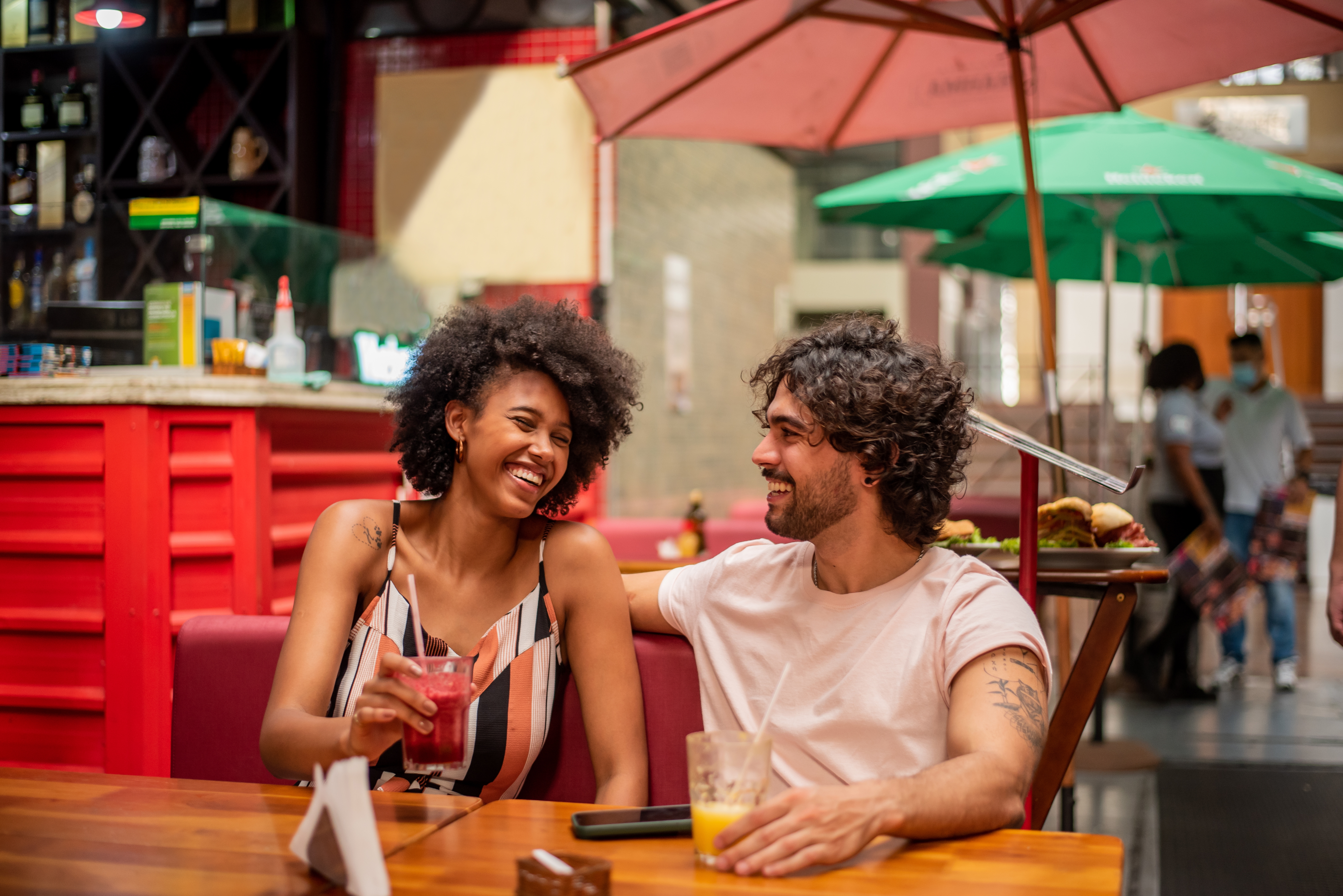 A couple smiling and chatting at an outdoor cafe, each holding drinks, suggesting a relaxed and happy atmosphere