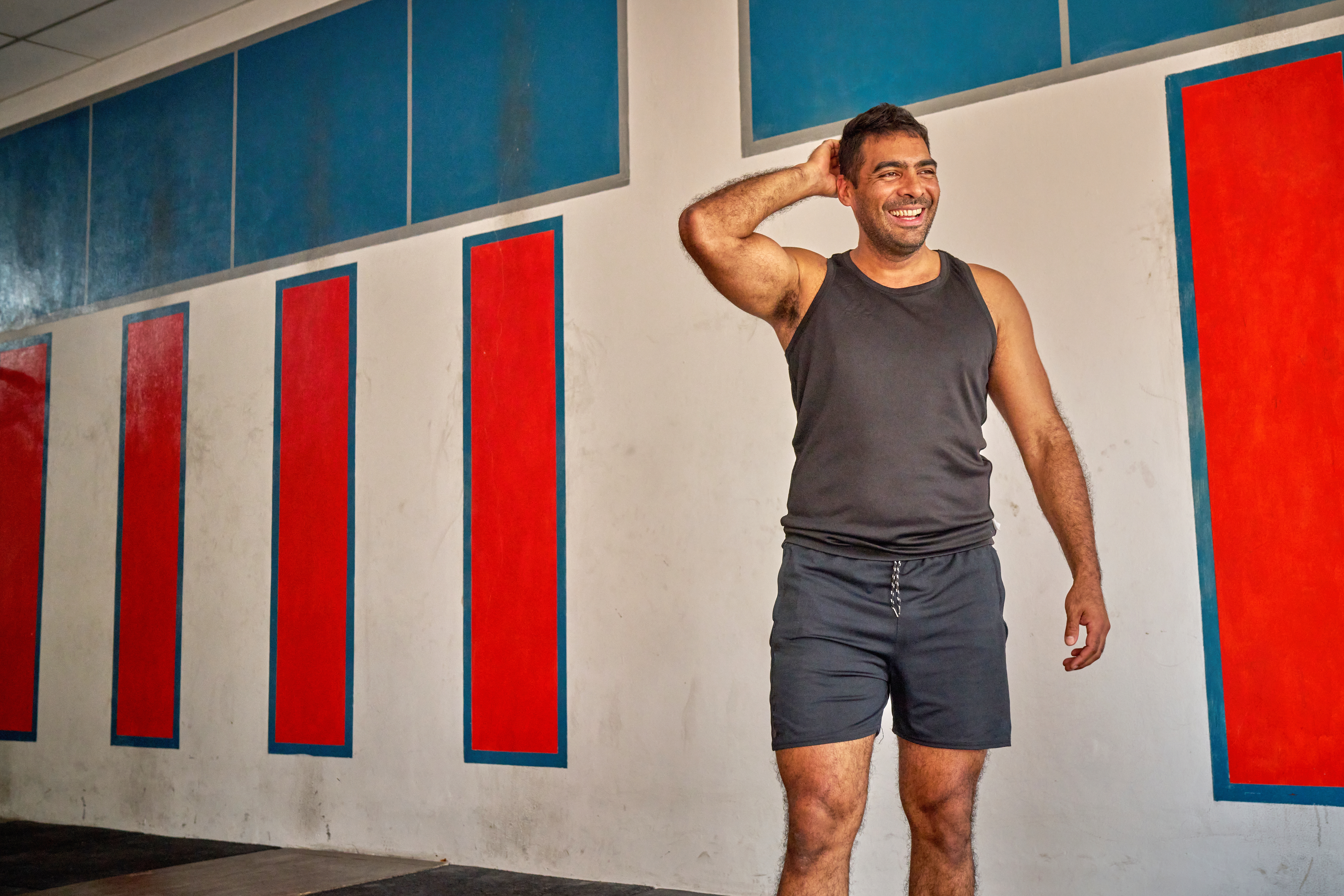 Man in tank top and shorts, smiling and posing against a wall with red and blue panels. Casual and relaxed atmosphere