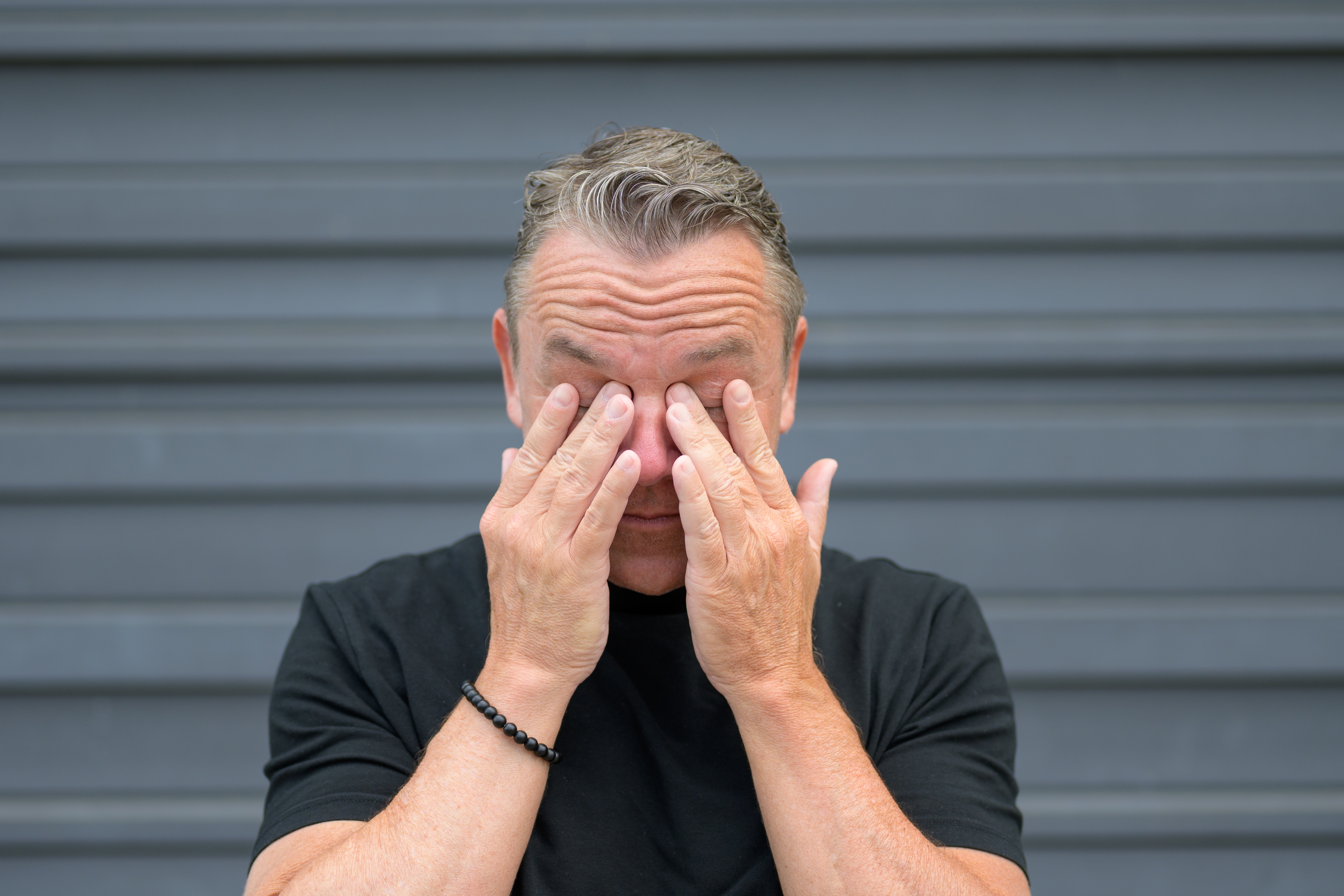 Man in black shirt rubs eyes in front of gray background, conveying emotion or fatigue