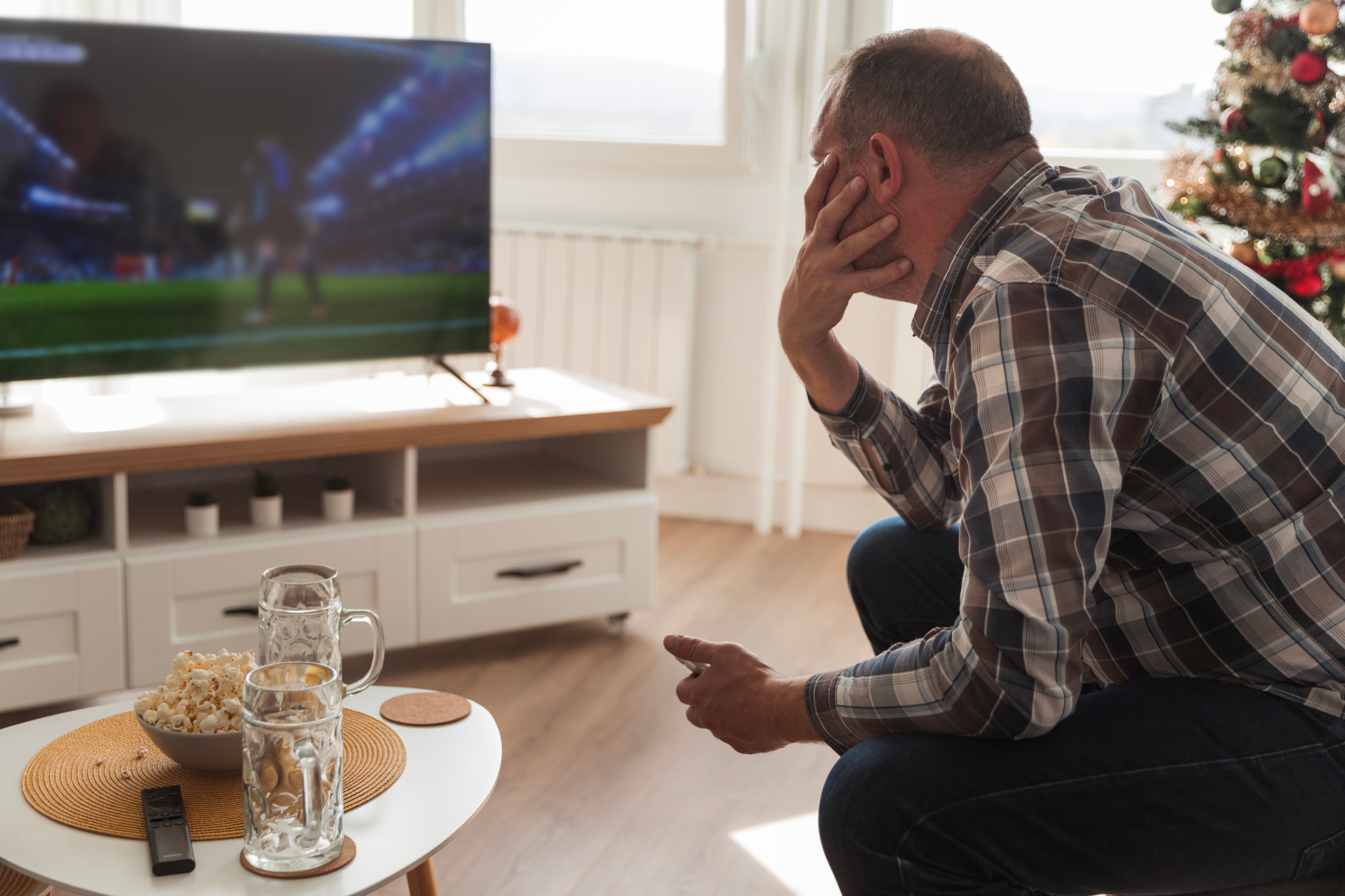 Man in plaid shirt watching sports on TV, sitting on a couch by a table with beer and popcorn, near a festively decorated Christmas tree
