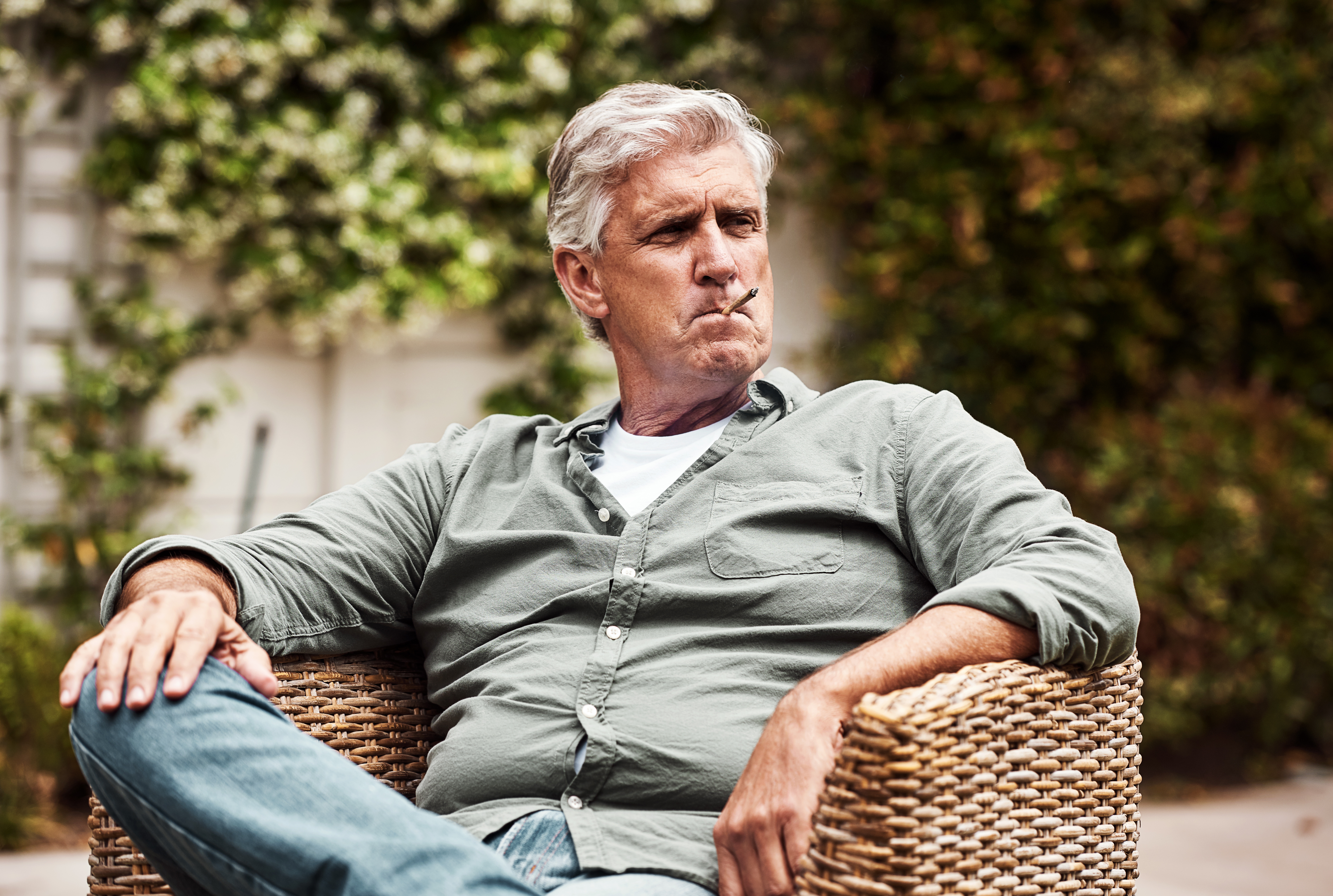 An older antheral   sits outdoors successful  a wicker chair, looking to the broadside  portion    holding a cigar successful  his mouth, appearing relaxed and thoughtful