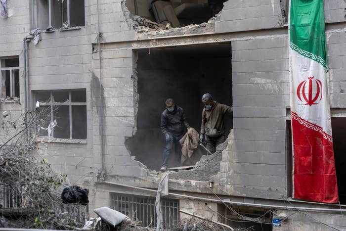 Damaged gathering  facade with 2  individuals clearing debris; an Iranian emblem  hangs alongside
