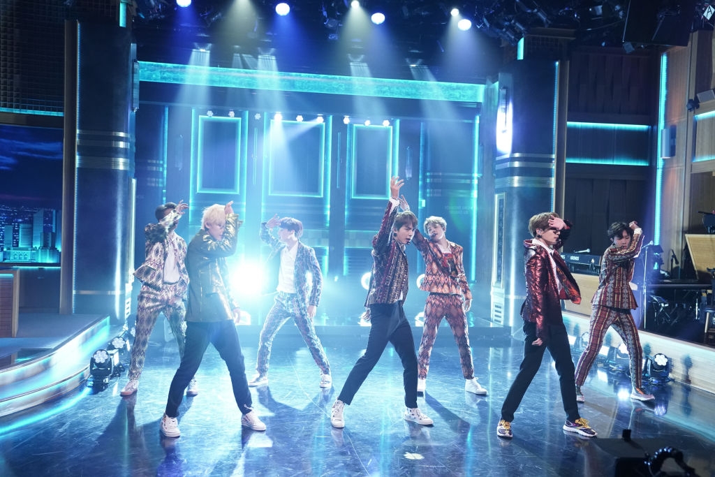A group of musicians performs energetically on a stage with dramatic lighting, wearing stylish and coordinated outfits