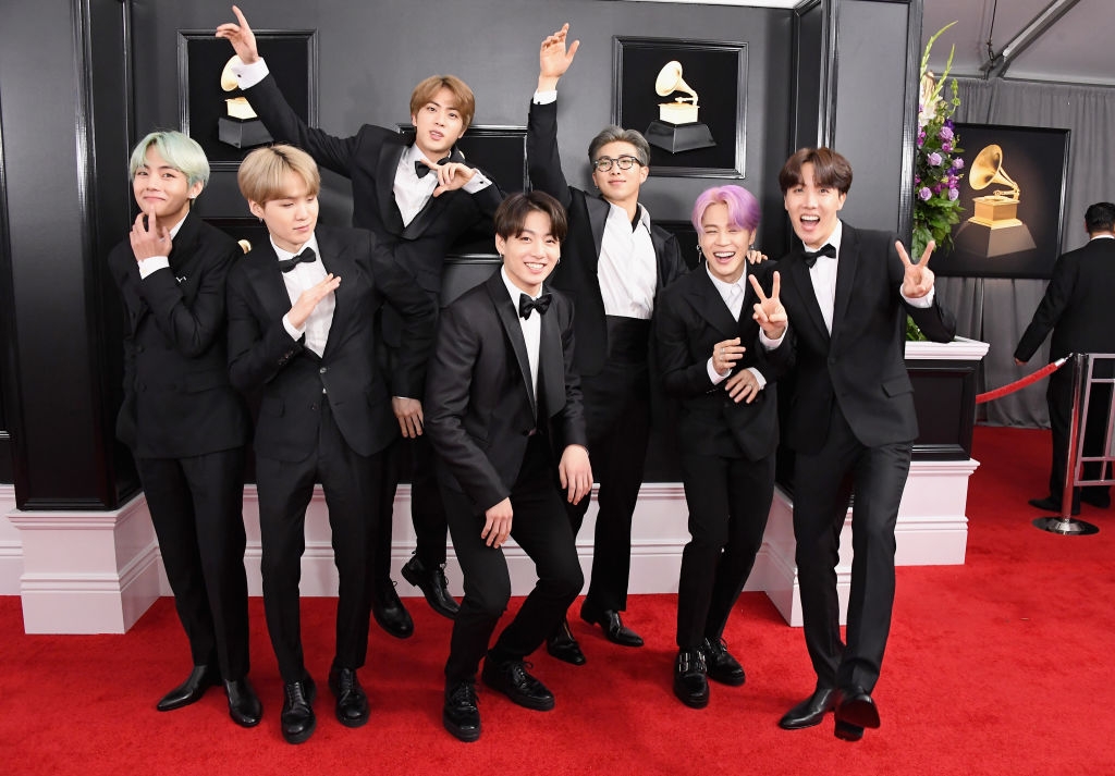 A group of seven people poses on the red carpet in tuxedos, celebrating joyfully with various playful hand gestures