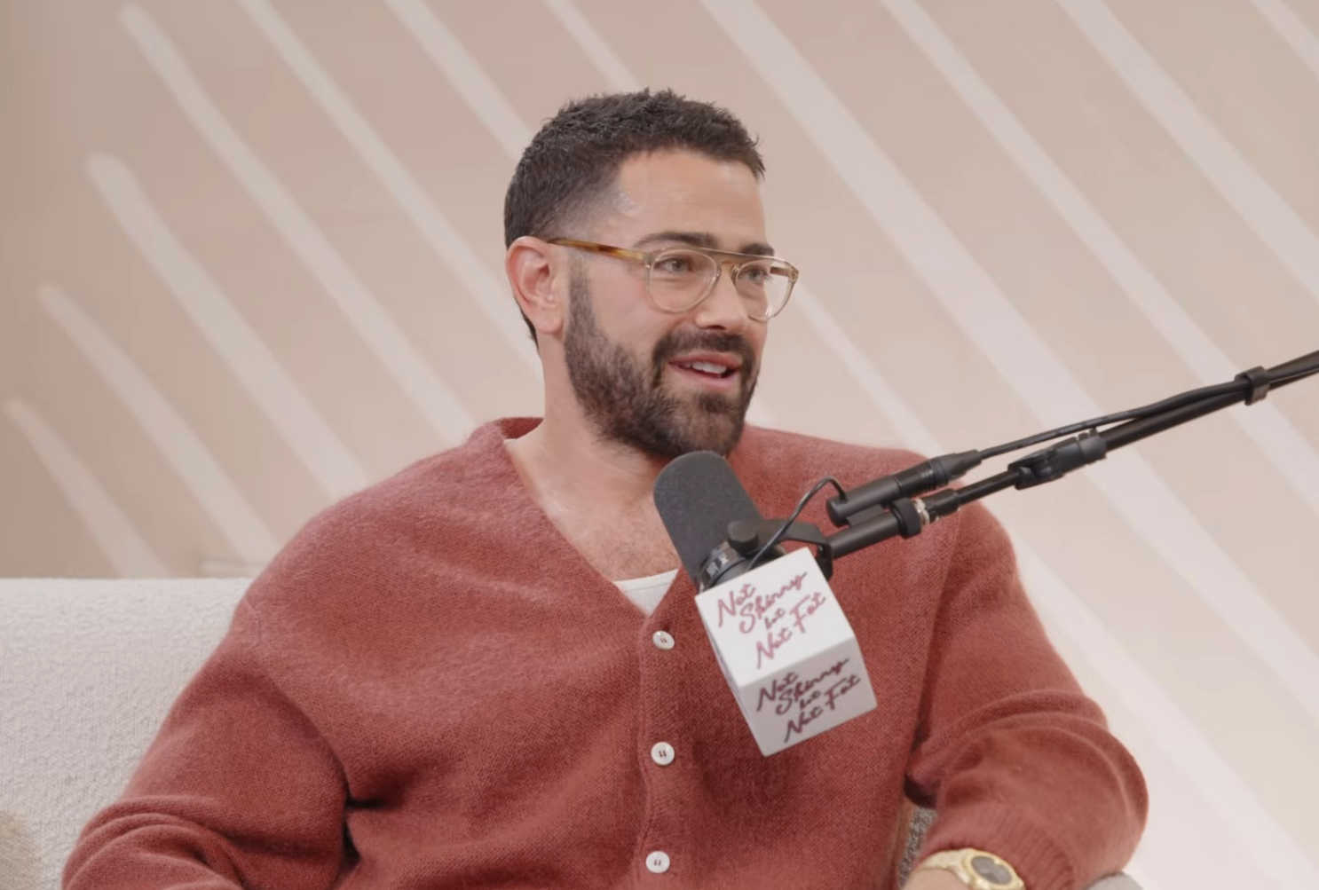 Person speaking into a microphone during a podcast, wearing a cardigan, glasses, and a watch, seated connected  a sofa successful  a workplace  setting