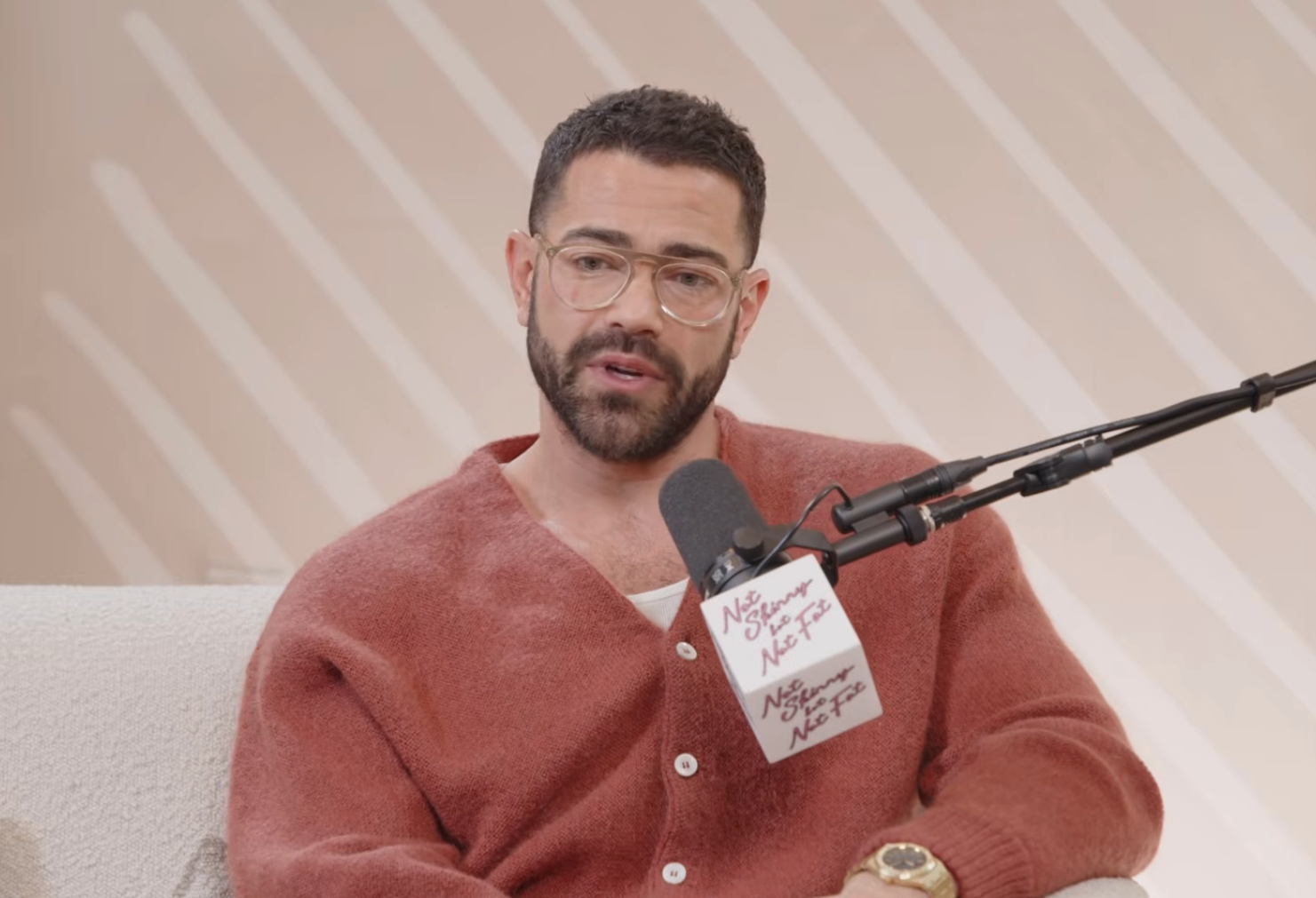 Person with beard and glasses speaking into a podcast microphone, wearing a casual cardigan and seated connected  a couch