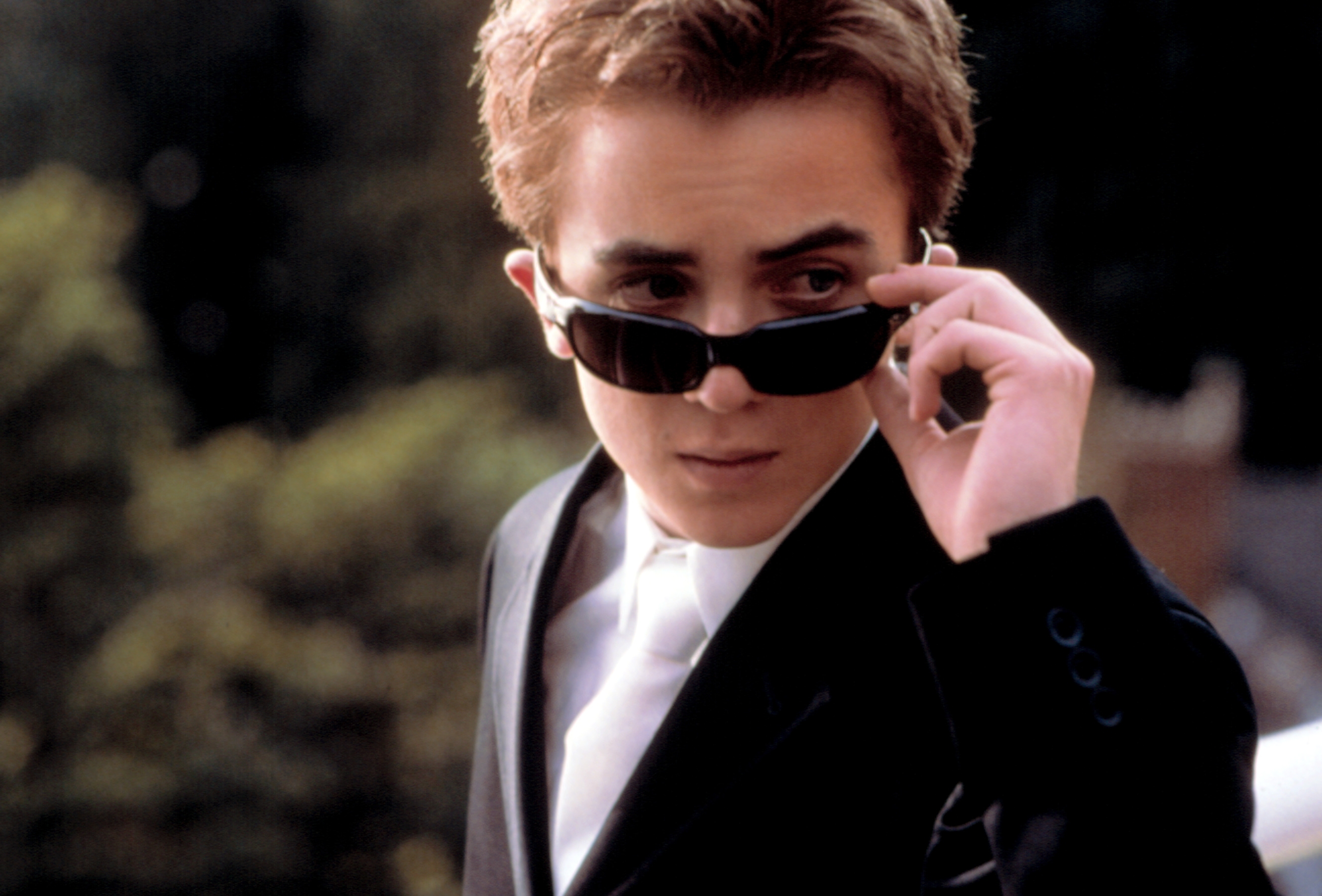 A idiosyncratic   successful  a sleek suit   adjusts their sunglasses with a assured  expression. Background is blurred greenery