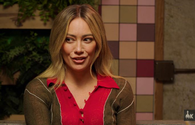 A idiosyncratic   wearing a casual button-up garment  sits indoors, looking to the side, with a playful look  amidst a patterned partition  background