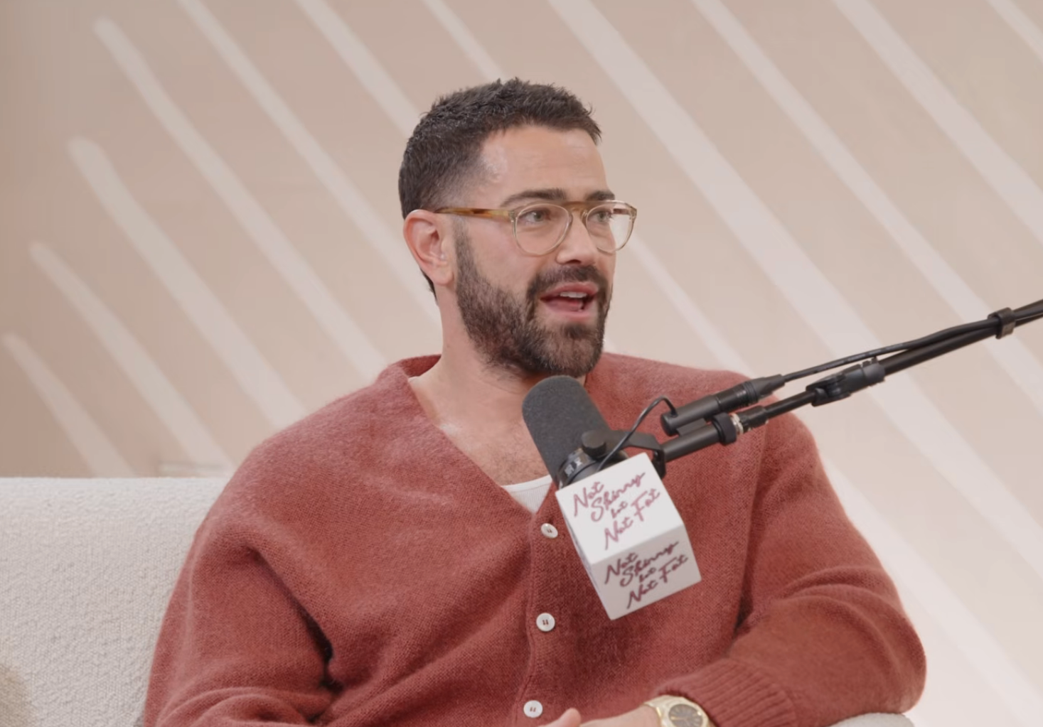 A idiosyncratic   wearing glasses and a cardigan speaks into a microphone during a podcast recording