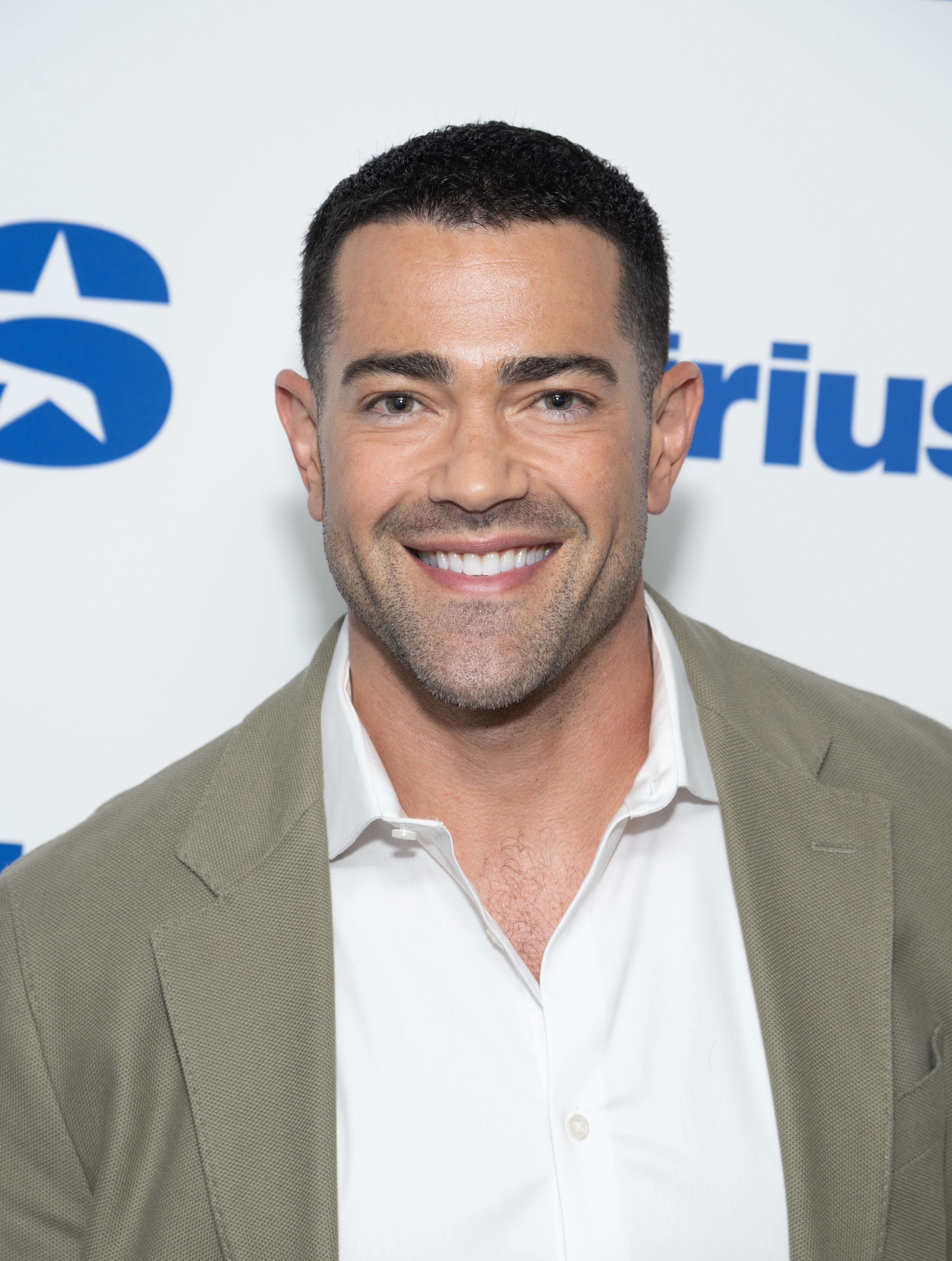 Man successful  a suit   and open-collared garment  smiles astatine  an event. Steps and repetition  backdrop with institution  logos down  him