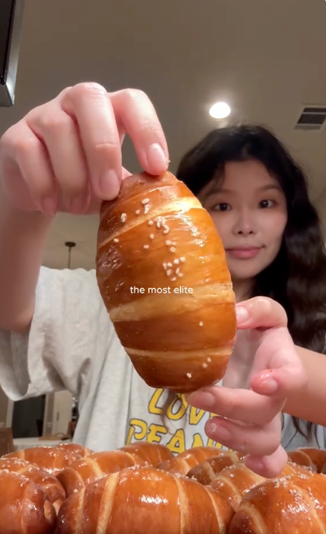 Person holding a freshly baked, glossy croissant with sesame seeds, surrounded by much  croissants. Text connected  representation  says, "the astir   elite."