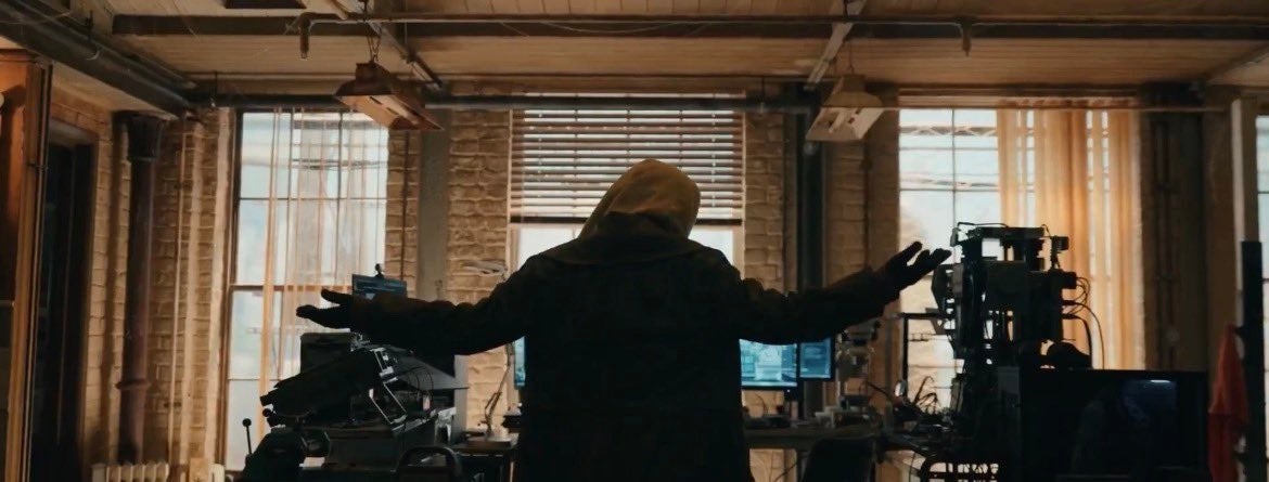 En person i en hettegenser står overfor flere dataskjermer i et svakt opplyst, teknologifylt rom, med armene utstrakt, noe som antyder et dramatisk eller anspent øyeblikk